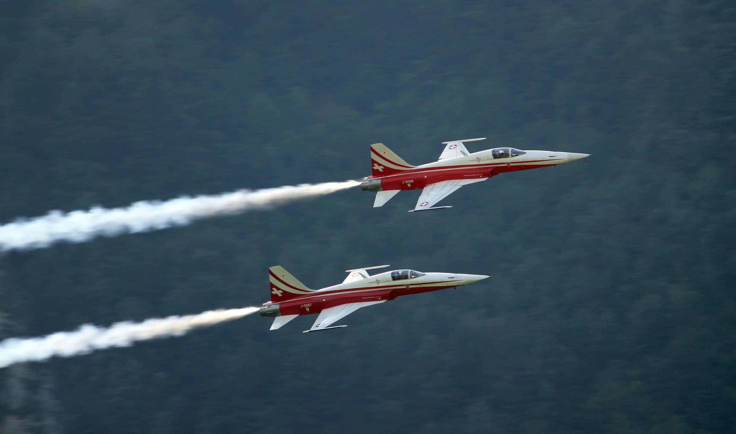 在小飛機中仰望天空 壁紙 — 兩架紅白相間的戰鬥機以緊密編隊飛越茂密的綠色森林，留下白色煙霧痕跡。