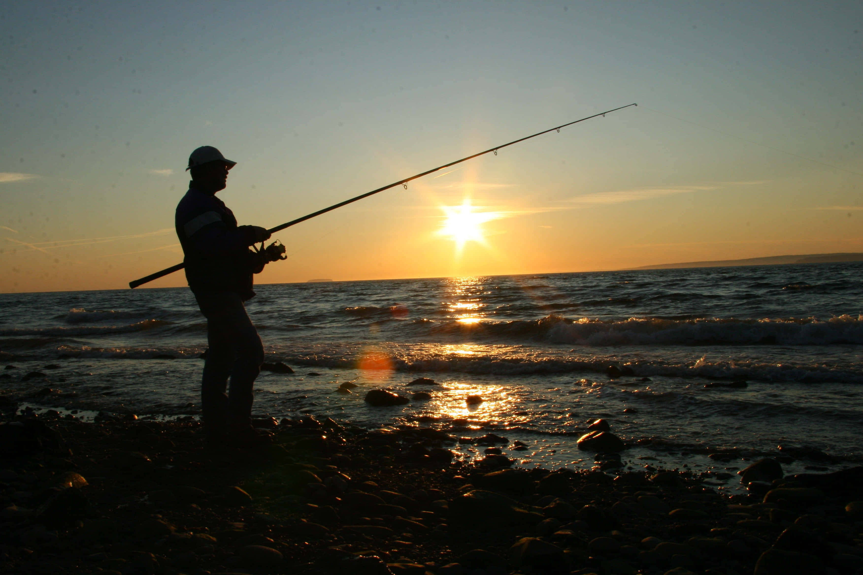 Fishing Rod 桌布 壁紙 — 在岩石海岸線上，夕陽下漁夫拋線的剪影，金色光芒反射在波浪上。