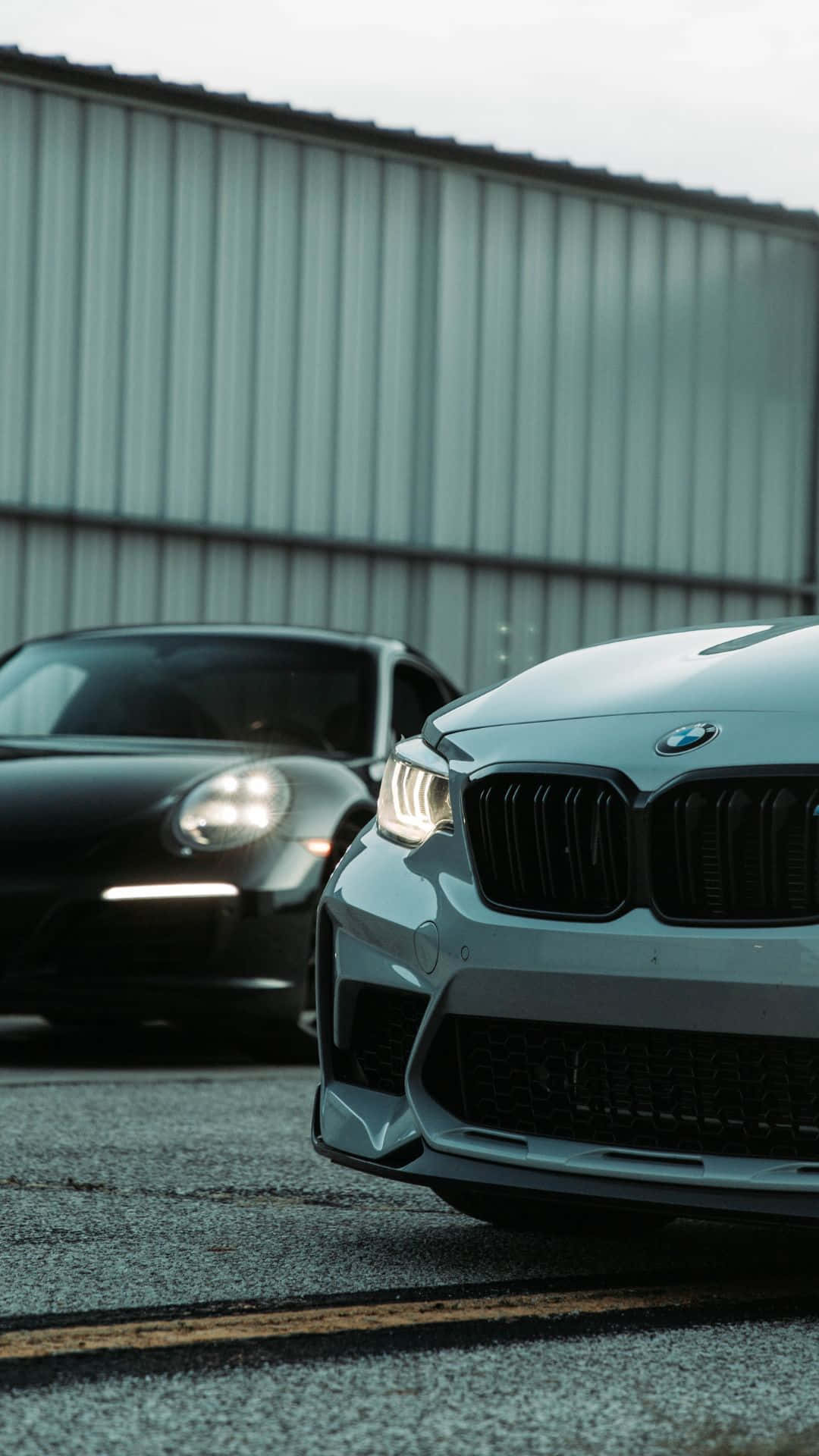在您的 BMW 中享受最新 Android 功能 壁紙 — 兩輛時尚跑車停在瀝青路上，背景是波紋金屬建築，天空多雲。
