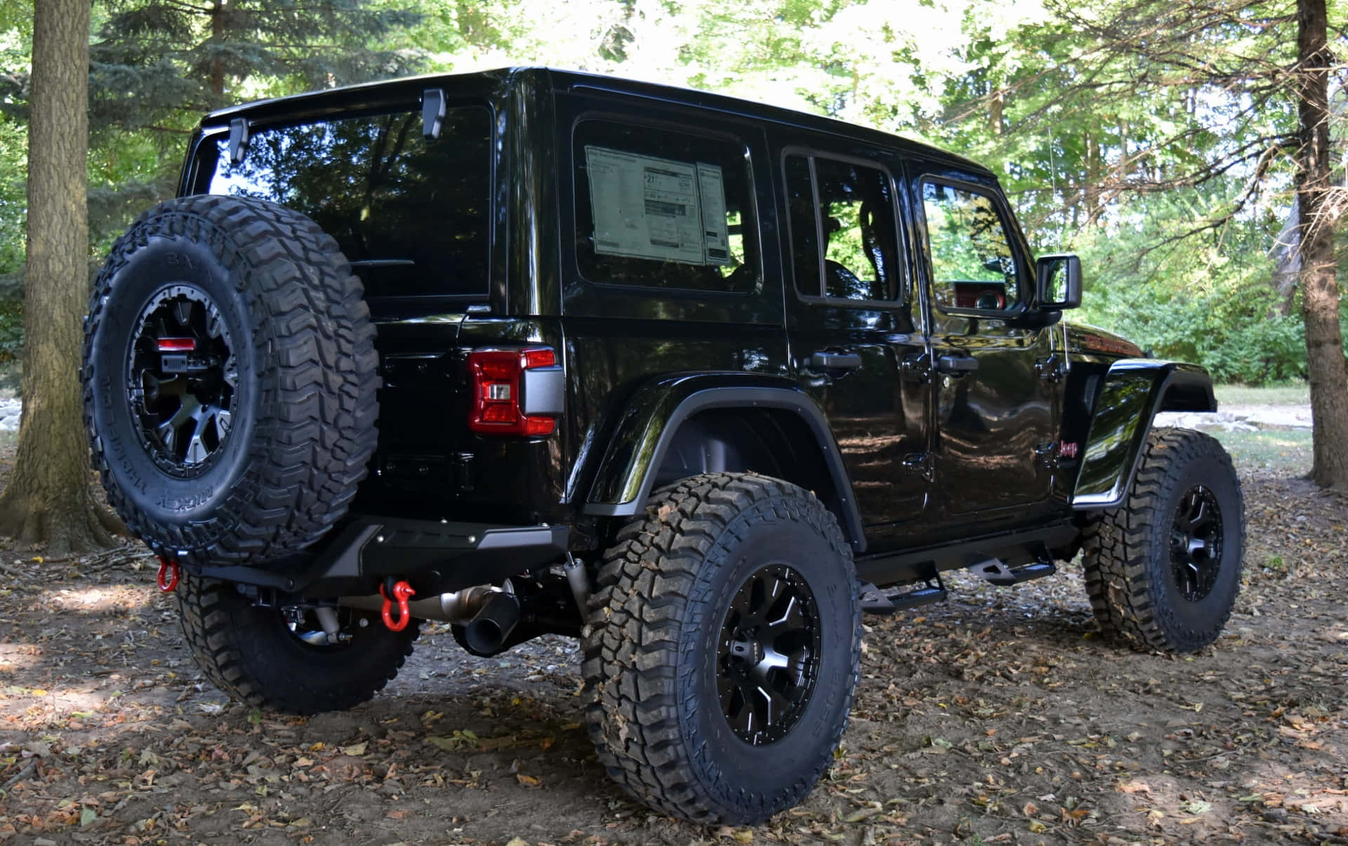 在您的Jeep Wrangler中展現個人風格 壁紙 — 一架亮黑色Jeep Wrangler配備超大輪胎，停泊在樹葉茂密的森林地面上，陽光斑駁灑落。