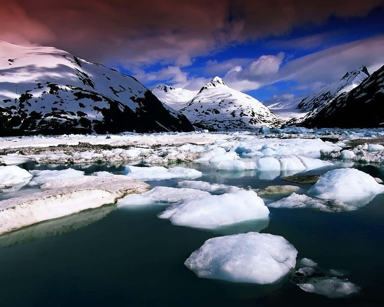Greenland 桌布 壁紙 — 在戲劇性的暮色天空下，冰川補給的河流中漂浮著冰山，背後是積雪覆蓋的山脈。