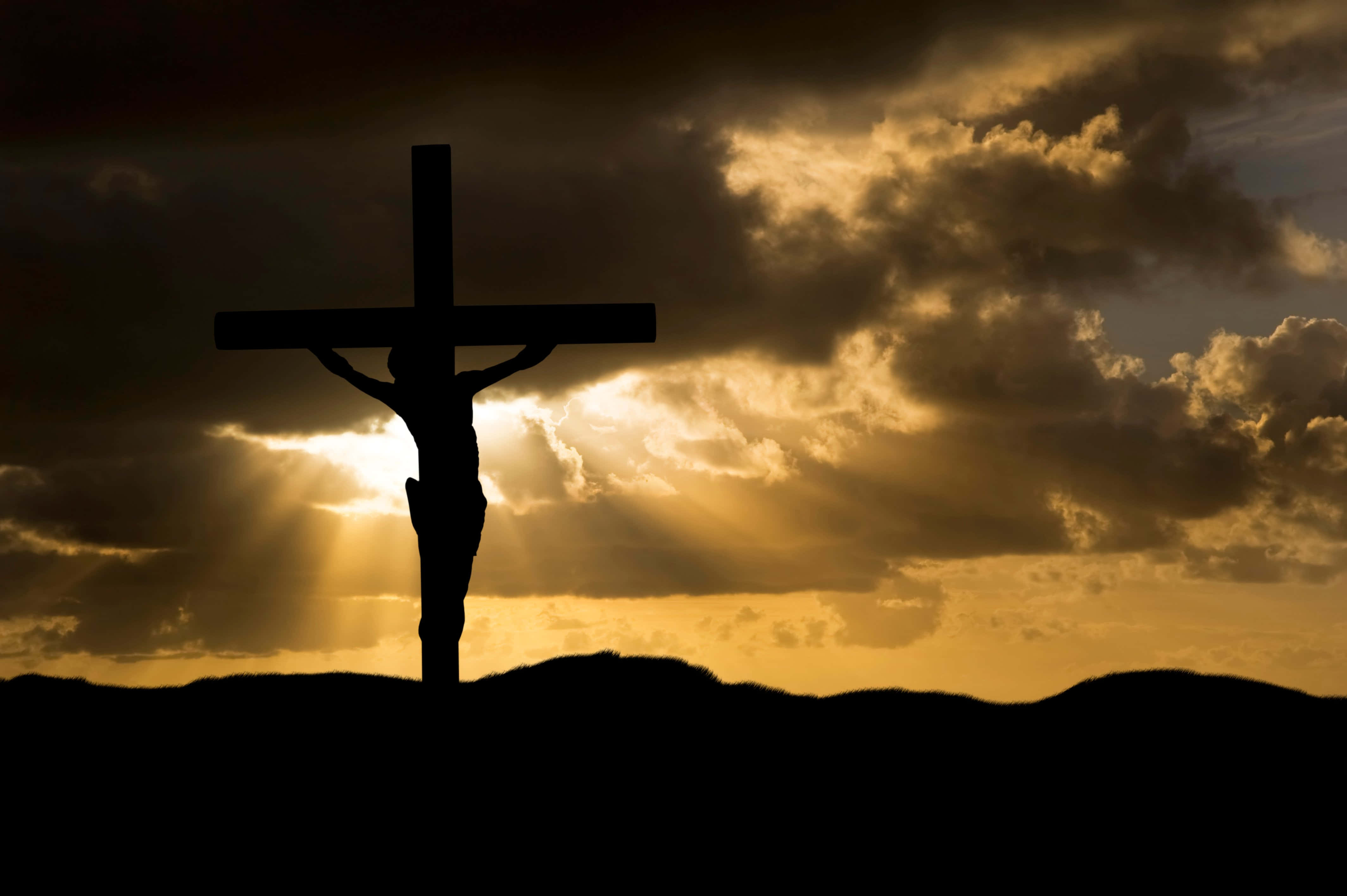 Jesus Crucifixion 桌布 壁紙 — 在戲劇性的金色日落背景下，十字架受難的剪影，耀眼的光芒穿透黑暗的雲層。