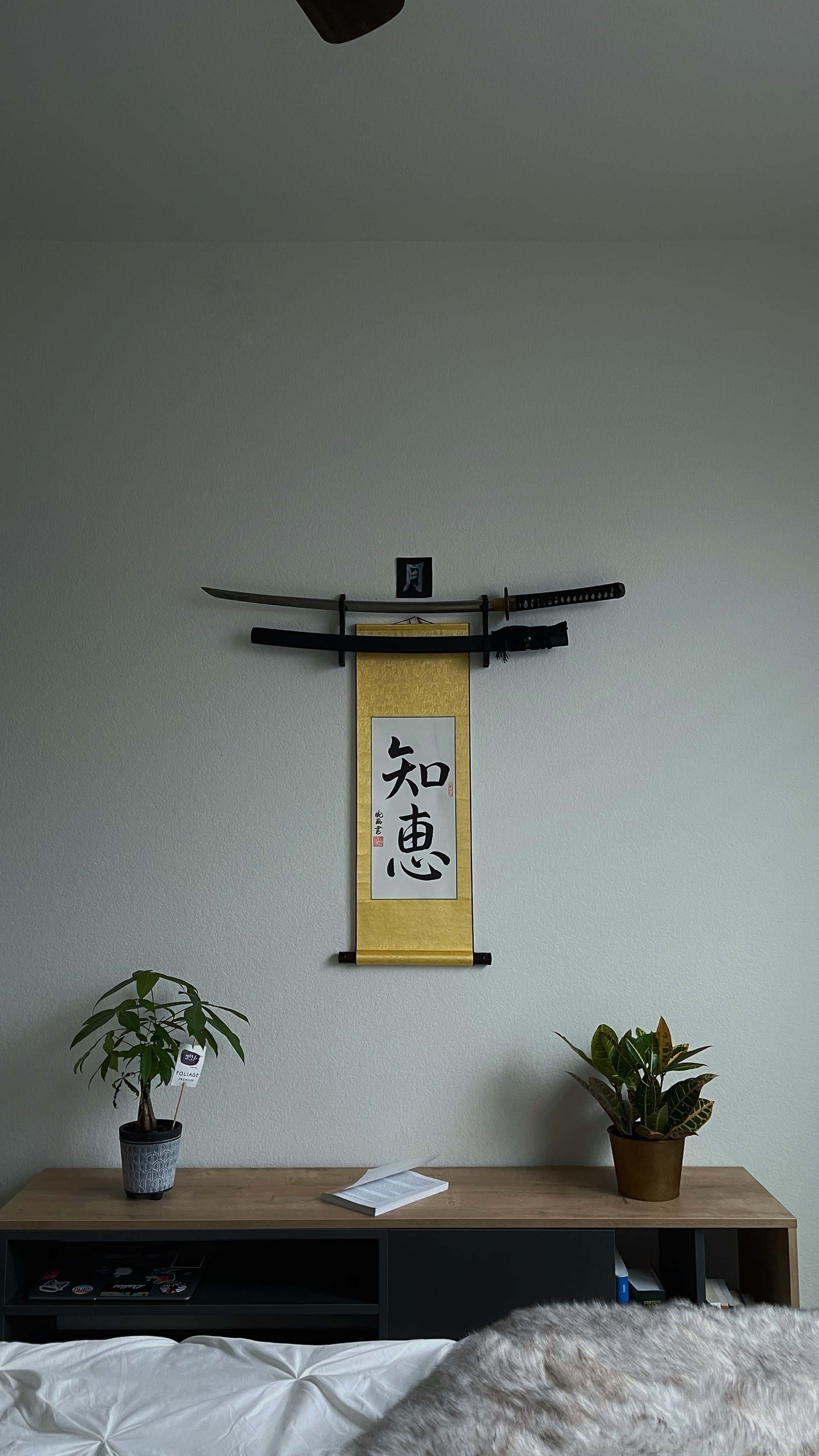 在日式白雅致中尋得寧靜 壁紙 — 簡約風格的臥室牆壁上展示兩把劍，上方懸掛著一幅日本書法卷軸，兩側擺放盆栽植物。