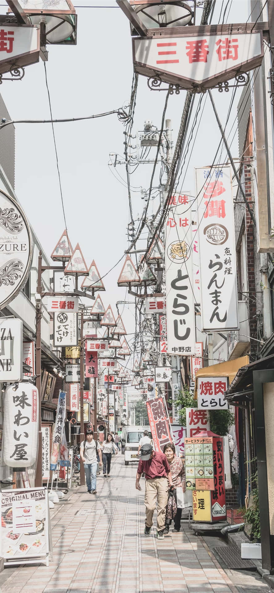 行人沿著一條狹窄的日本街道行走，街道兩旁掛滿色彩繽紛的招牌。