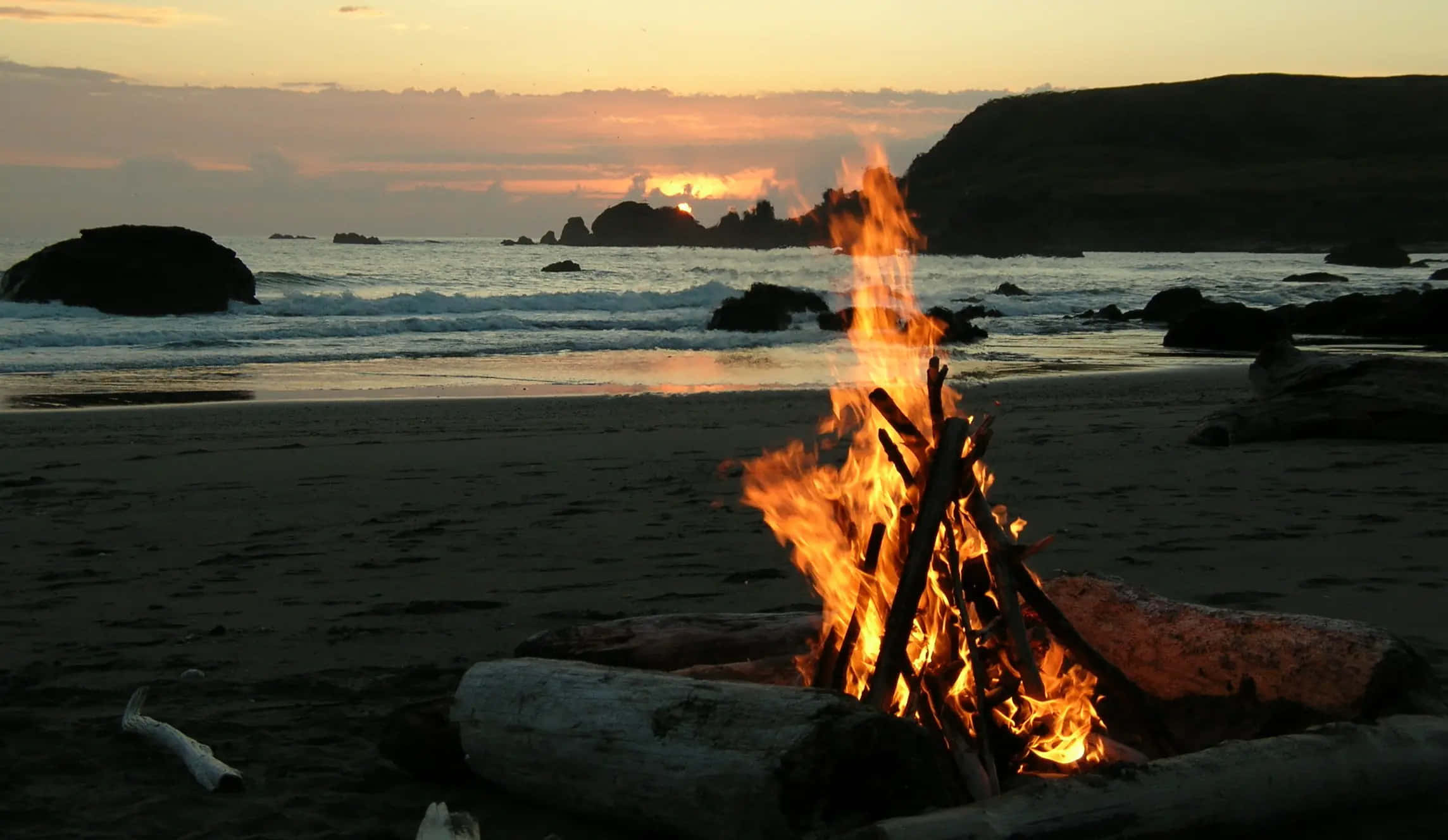 Beach Bonfire 桌布 壁紙 — 在日落時分，一個熊熊燃燒的營火在黑暗的沙灘上明亮地燃燒，海浪拍打著遠處的岩石和山丘。