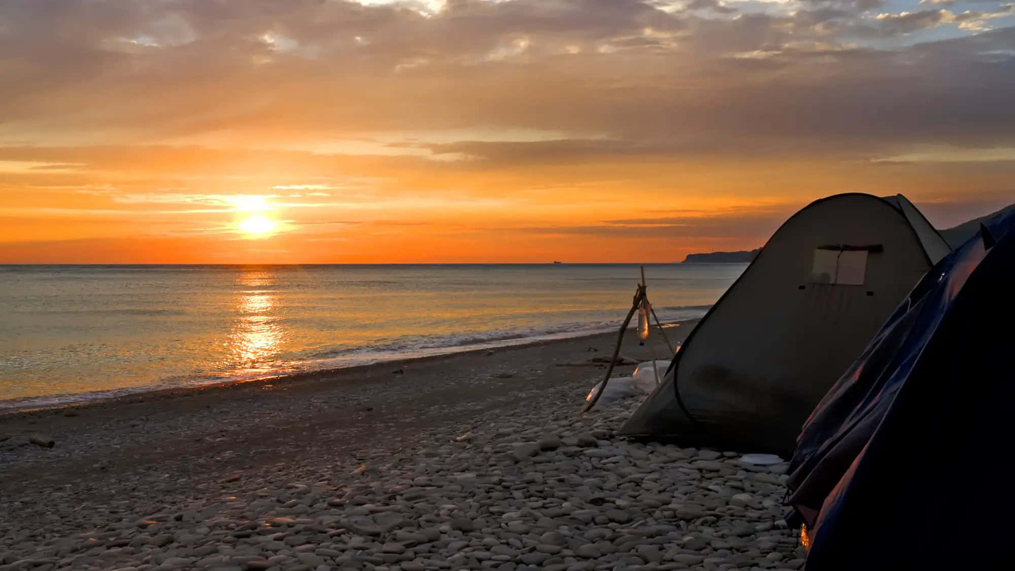 Beach Camping 桌布 壁紙 — 在日落時分，卵石海灘上搭起的帳篷，金色陽光映照在平靜的海面上。