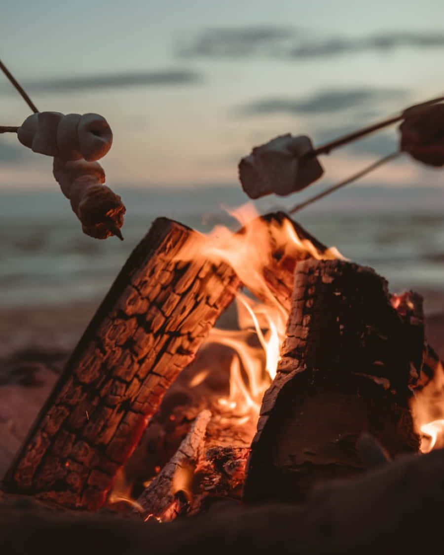 Beach Bonfire 桌布 壁紙 — 在日落時分，沙灘營火噼啪作響，烤著棉花糖，溫暖的橙色火焰和柔和的天色。
