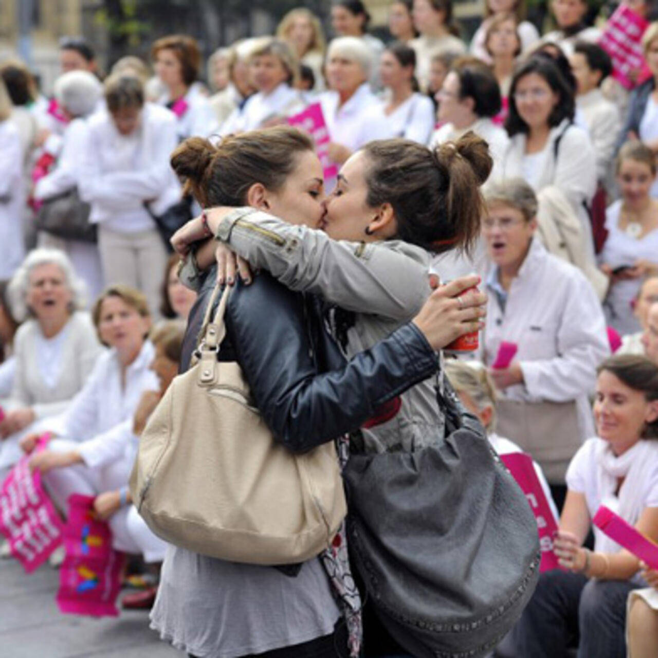 Girls Kissing at Protest 壁紙 — 在日間抗議活動中，兩名女性在舉著粉紅色標語牌的人群面前熱情擁抱和親吻。