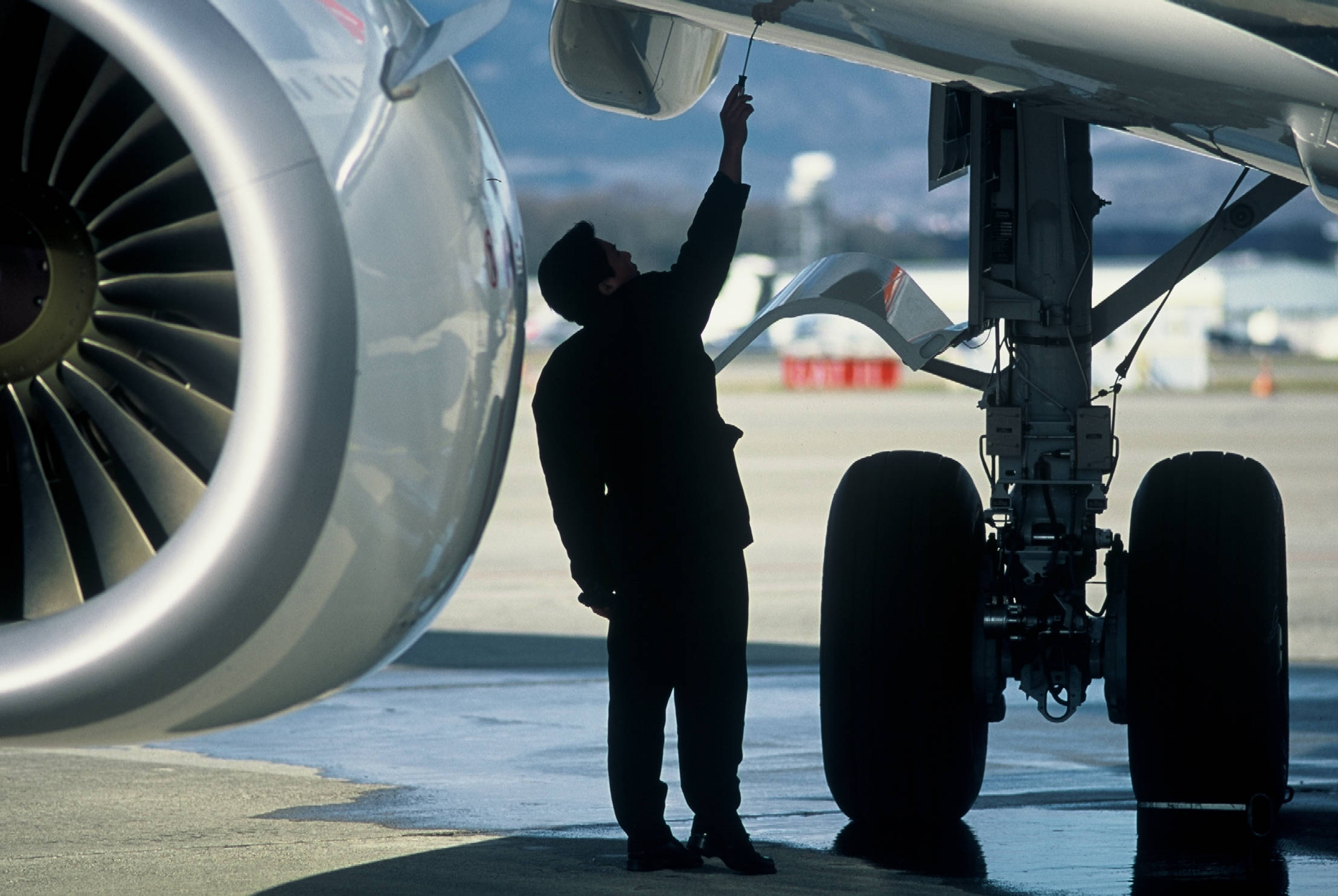 飛機 桌布 壁紙 — 在明亮日光下，機械師在濕滑停機坪上檢查飛機引擎的剪影。