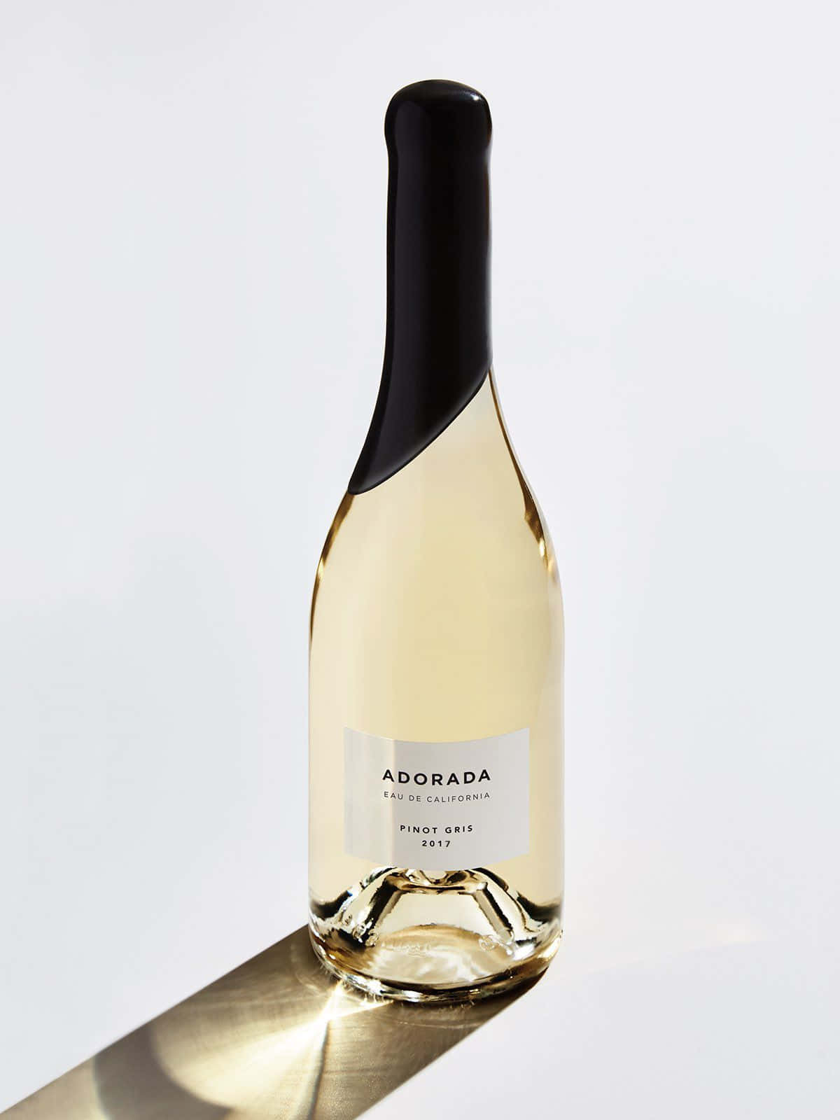 White Wine 桌布 壁紙 — 在明亮的攝影棚燈光下，一瓶流線型的Adorada Pinot Gris葡萄酒在簡約的白色表面上投下銳利的陰影。