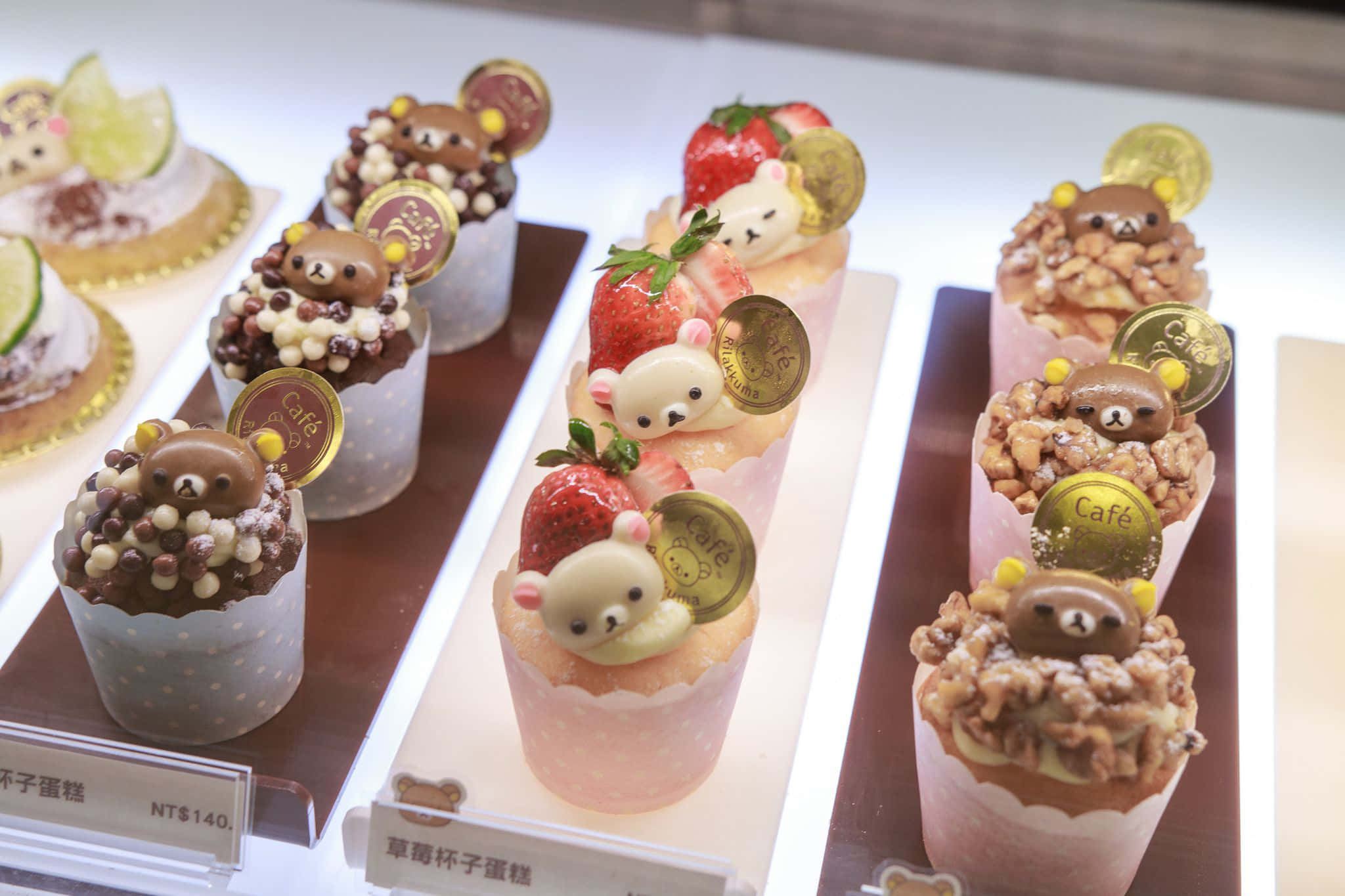 Kawaii Dessert 桌布 壁紙 — 在明亮的烘焙展示櫃中，擺放著可愛的熊主題杯子蛋糕，上面裝飾著草莓、堅果和金幣。