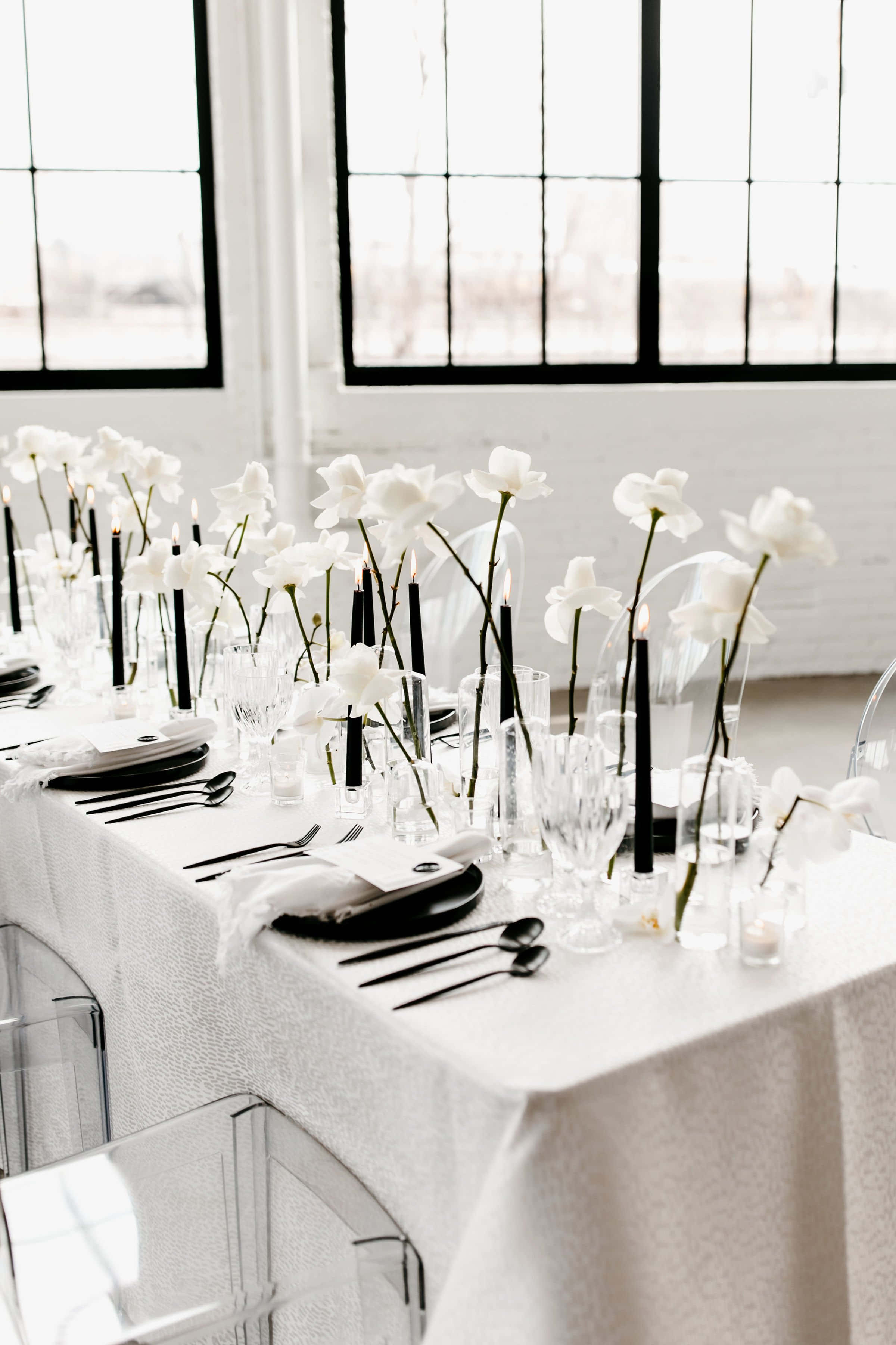Black And White Wedding 桌布 壁紙 — 在明亮的窗光下，優雅的黑白餐桌佈置，配有白色蘭花、高黑色蠟燭和透明玻璃器皿。