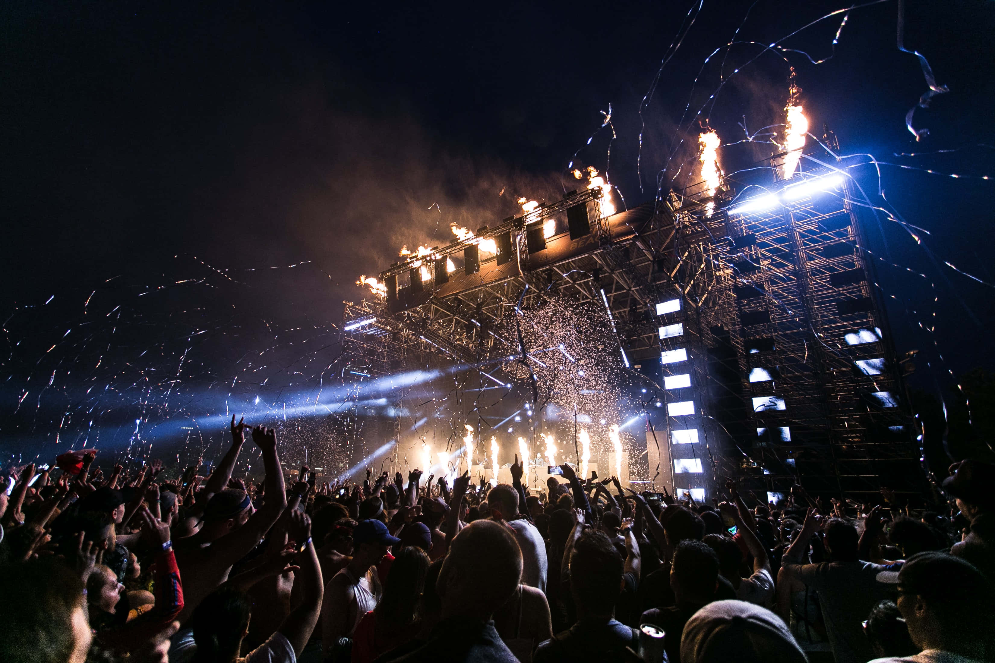 music festival 桌布 壁紙 — 在明亮的聚光燈下，夜間節日中的人群歡呼，舞台上有熾熱的煙火和閃閃發光的五彩紙屑。
