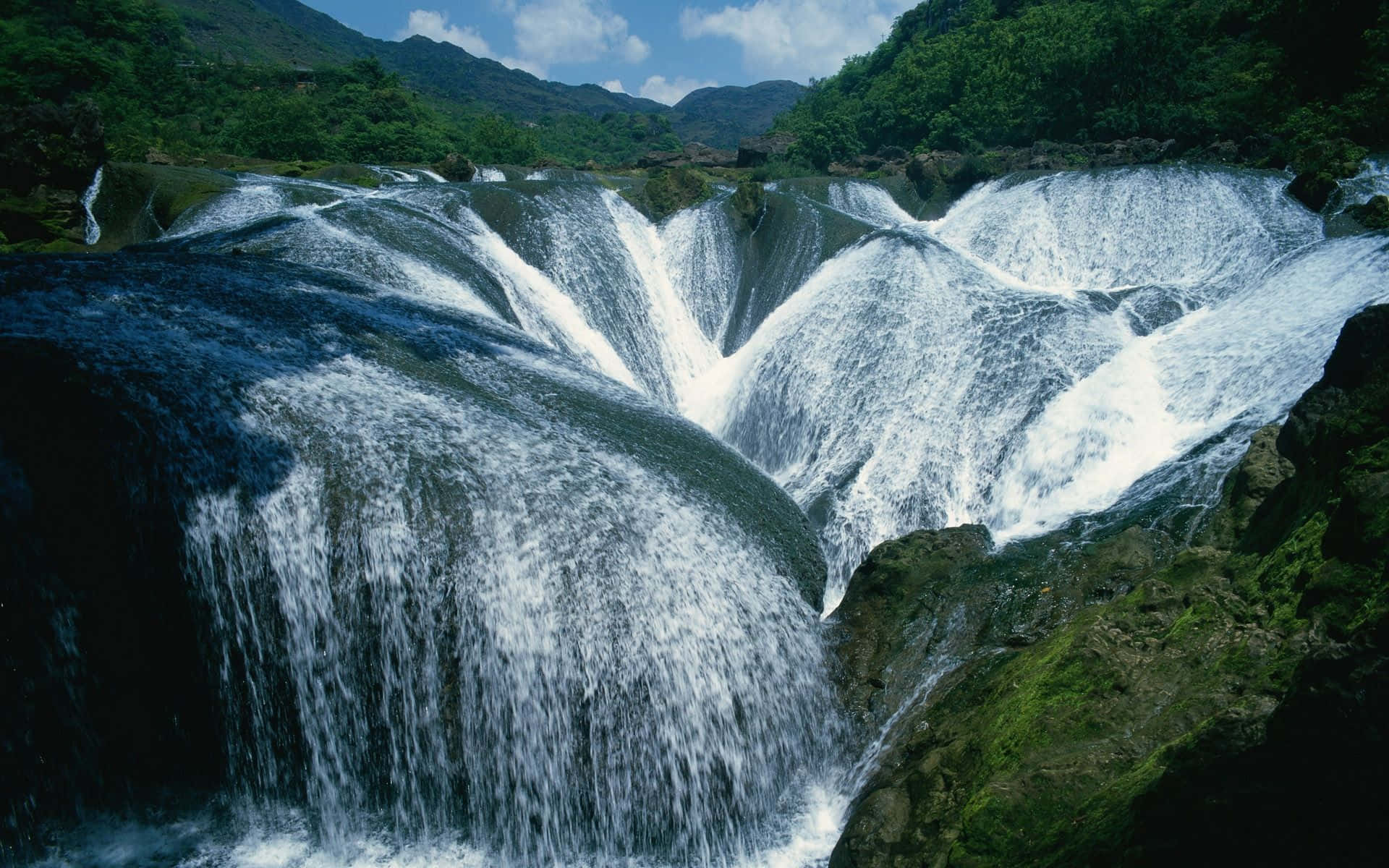 Waterfalls 桌布 壁紙 — 在明亮的藍天下，一個強大的多層瀑布從長滿苔蘚的岩石上傾瀉而下，流入鬱鬱蔥蔥的綠色山谷。