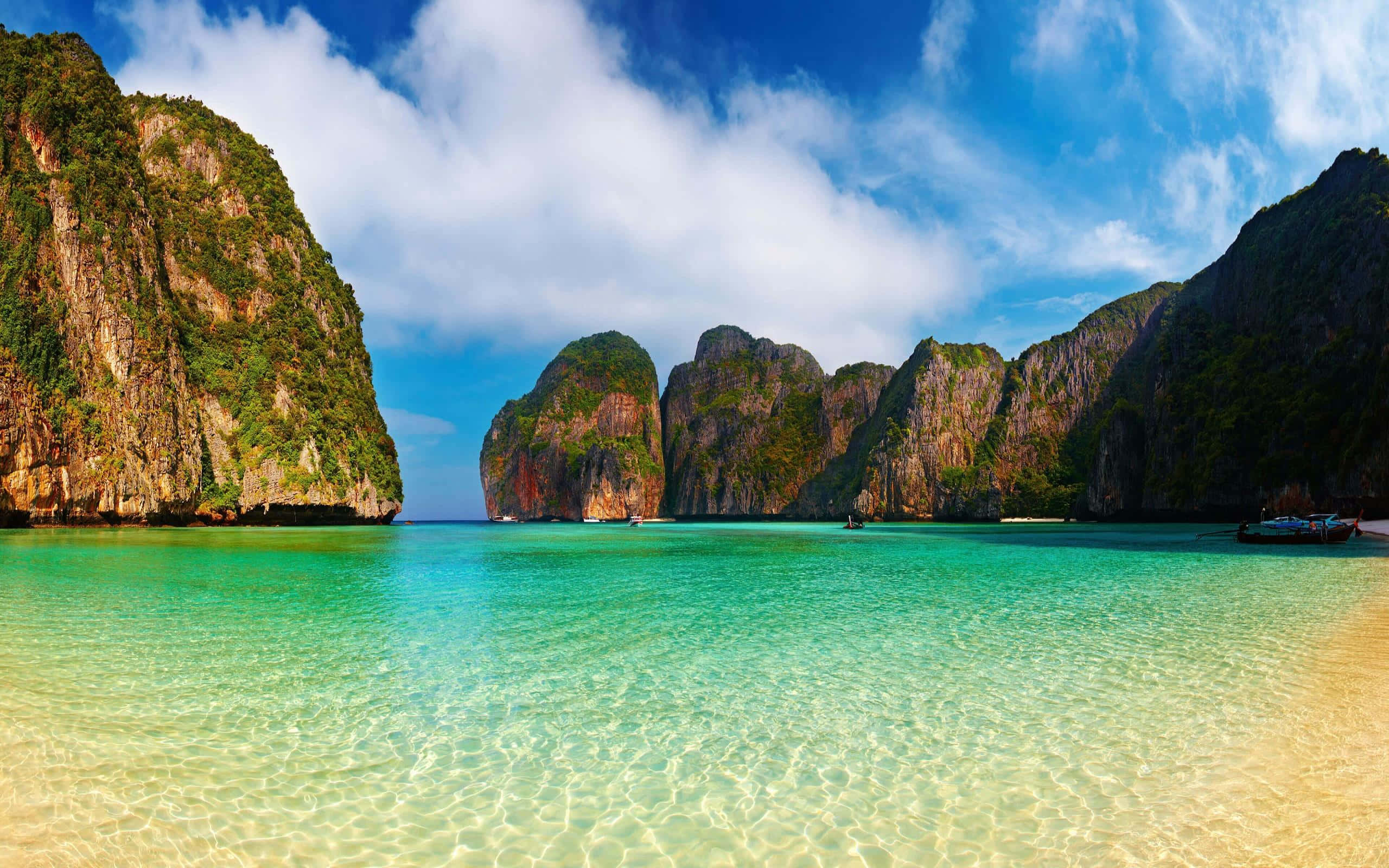 Thailand Beach 桌布 壁紙 — 在明亮的藍天下，翠綠的海水在一個被高聳的叢林覆蓋的石灰岩懸崖環繞的隱蔽熱帶海灣周圍輕輕蕩漾。