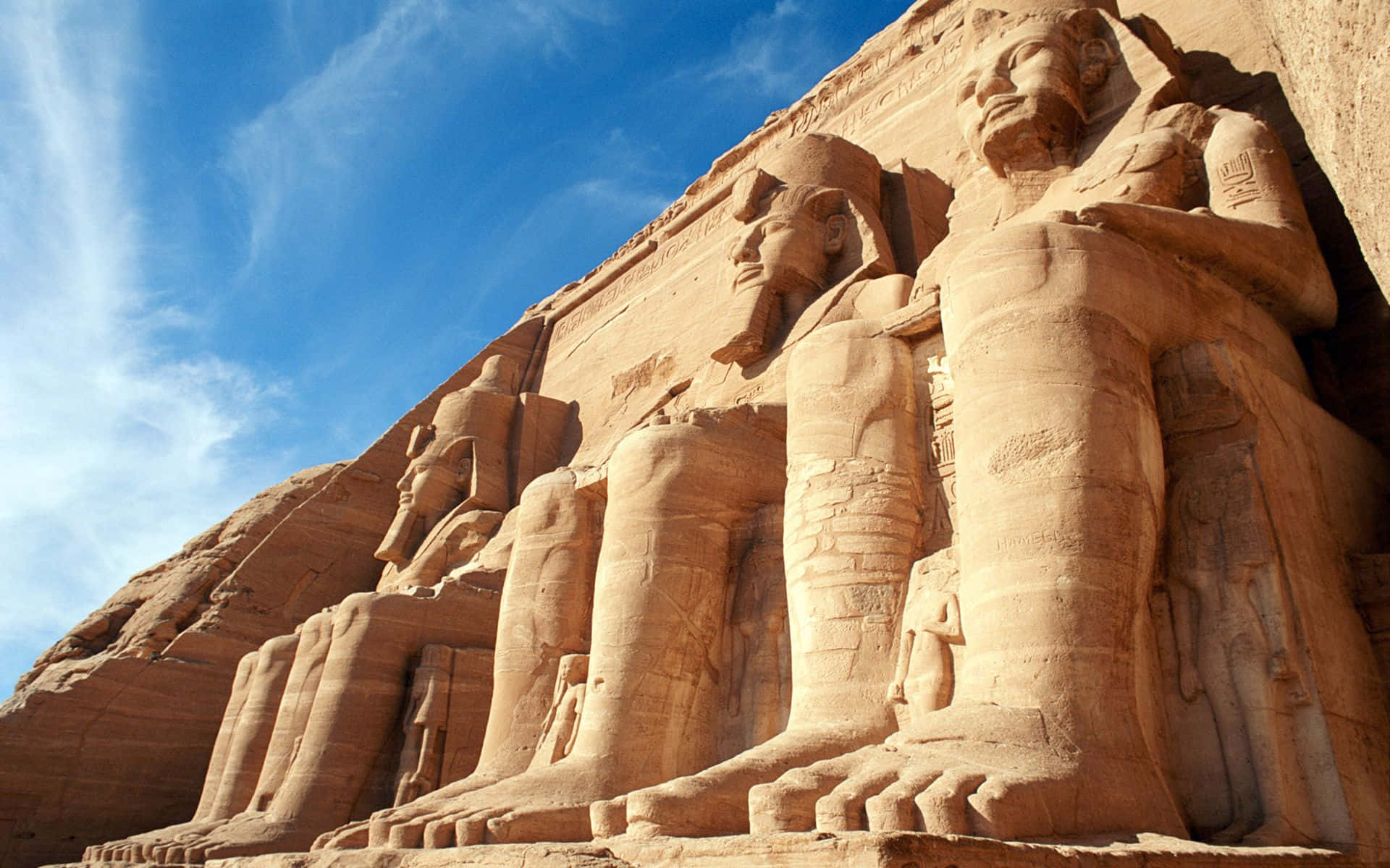 Egypt 桌布 壁紙 — 在明亮的藍天和薄雲下，砂岩懸崖上雕刻的巨大古埃及雕像。