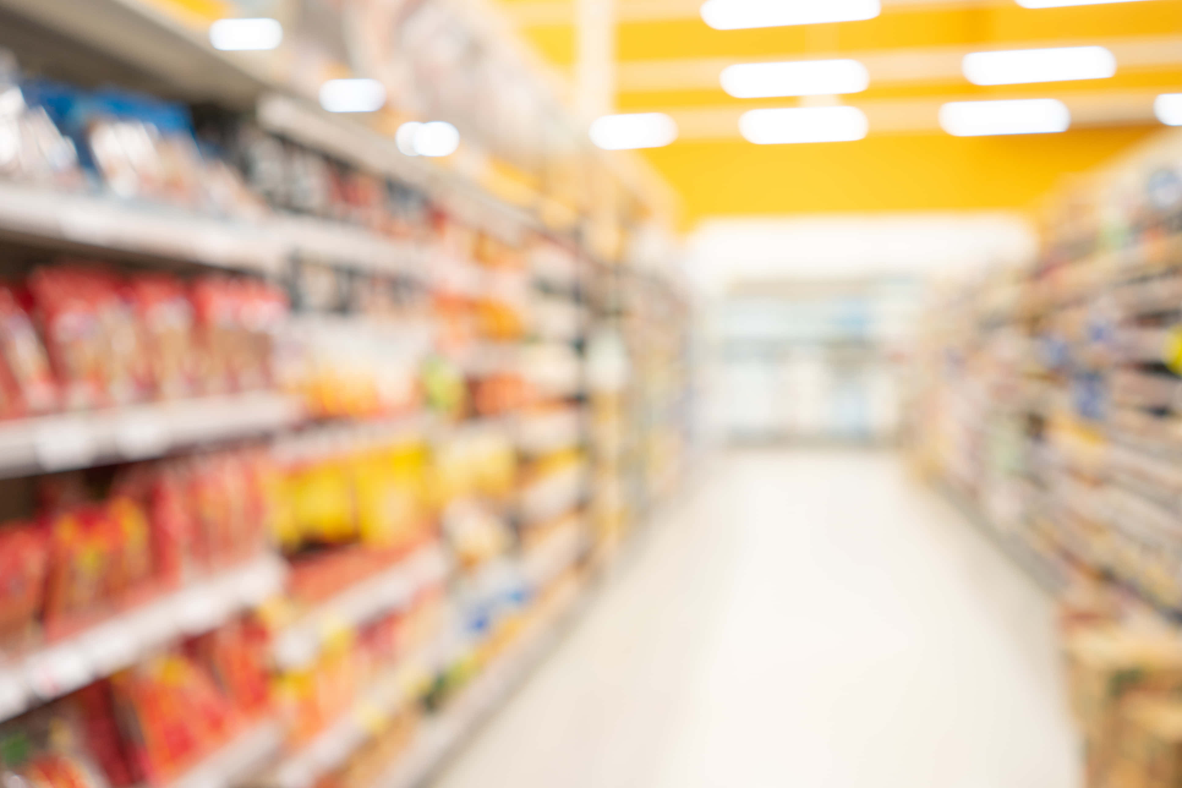 Grocery Store 桌布 壁紙 — 在明亮的黃色天花燈光下，模糊的超級市場通道，擺滿色彩繽紛的產品貨架。