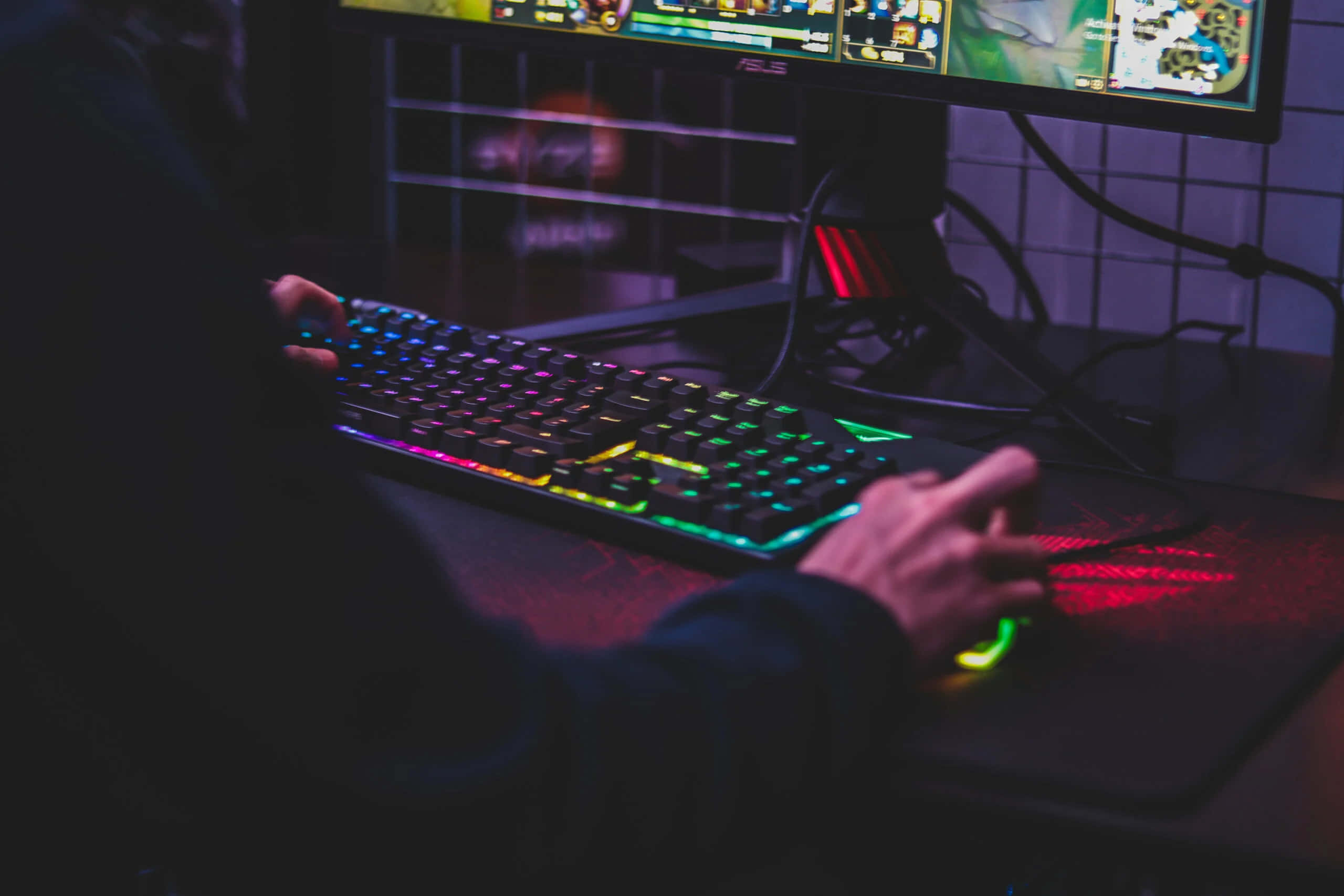 Gaming Mouse 桌布 壁紙 — 在昏暗的紫色房間燈光下，雙手在彩虹發光的遊戲鍵盤上打字，發光的顯示器顯示遊戲。