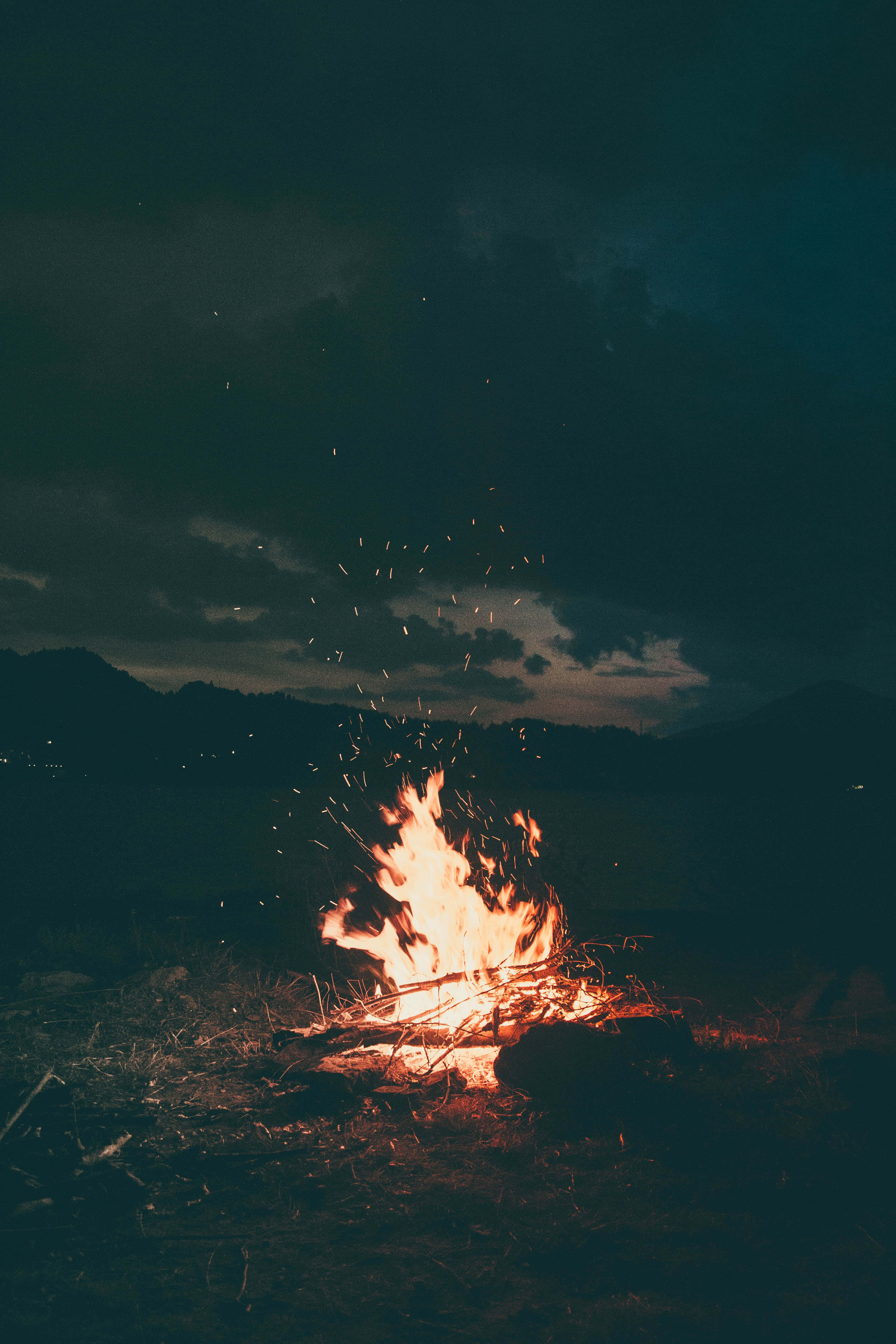 在晴朗夜晚篝火旁烤棉花糖 壁紙 — 在黑暗多雲的天空下，一個充滿活力的營火噼啪作響，發光的餘燼在夜間荒野環境中向上飛舞。