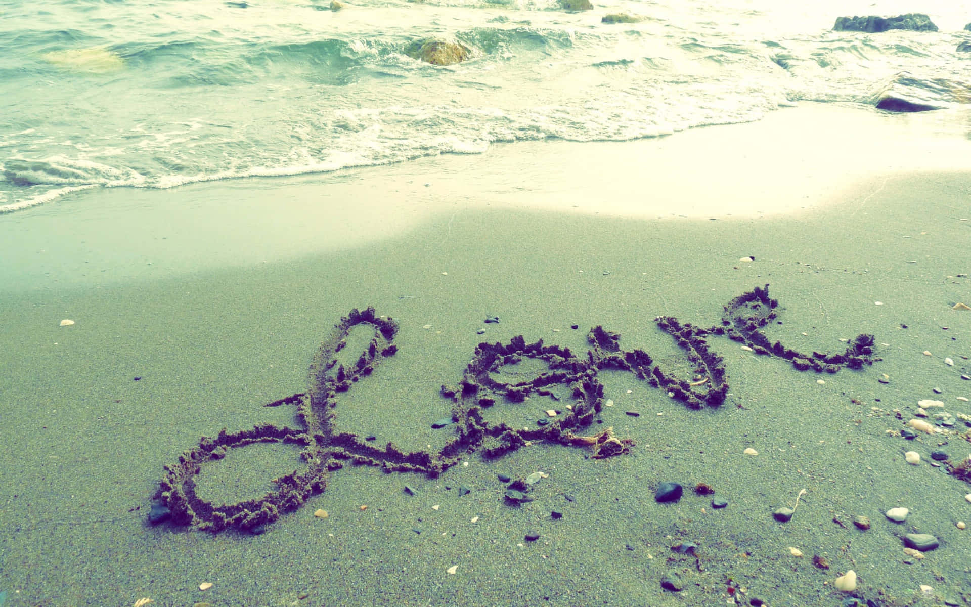 Beach Love 桌布 壁紙 — 在柔和日光下，溫柔的海浪拍打著濕沙，上面用深色海藻寫著「愛」這個字。
