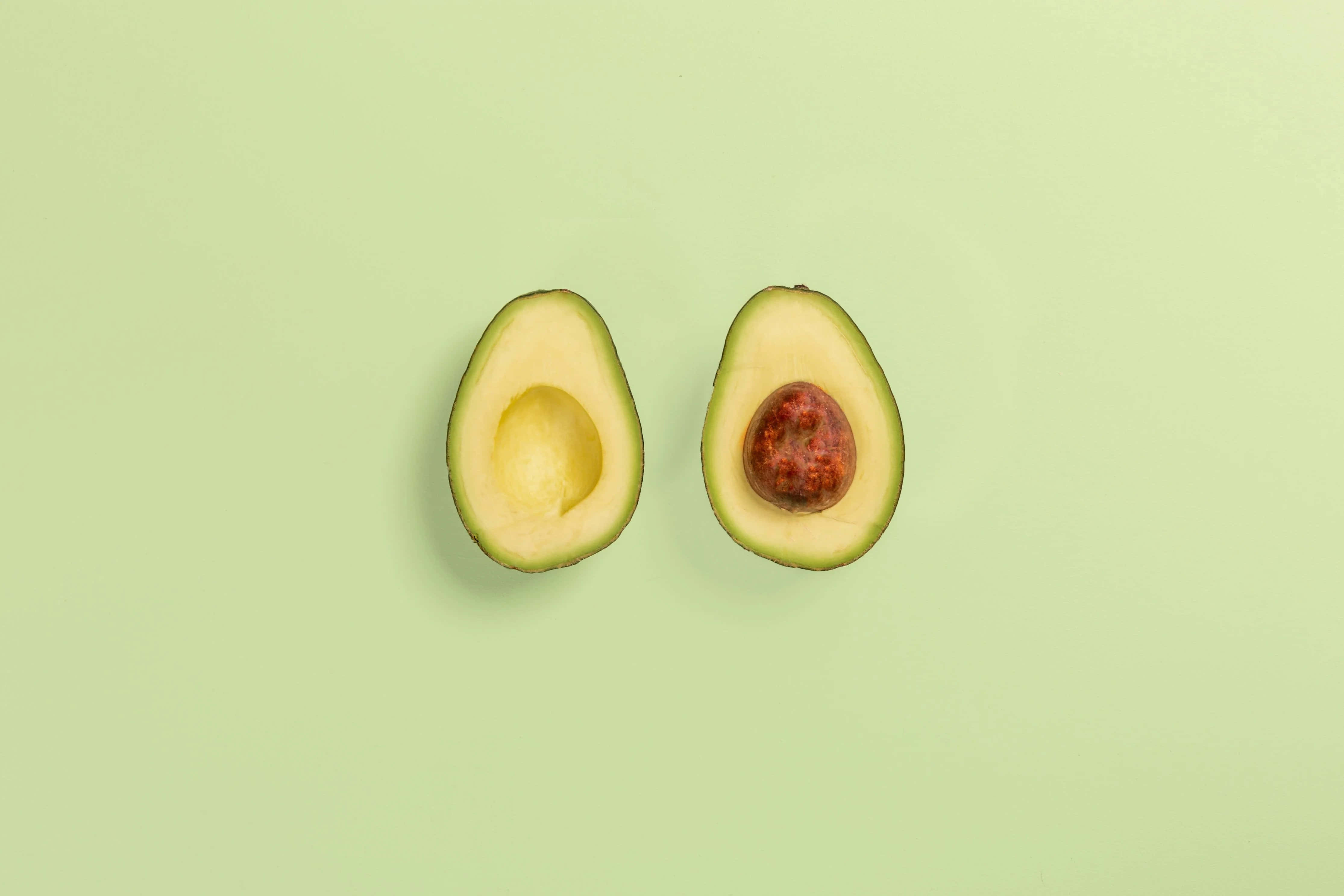 Avocado 桌布 壁紙 — 在柔和的薄荷綠色背景上，一個成熟的牛油果被切成兩半，露出奶油狀的果肉和棕色的果核。
