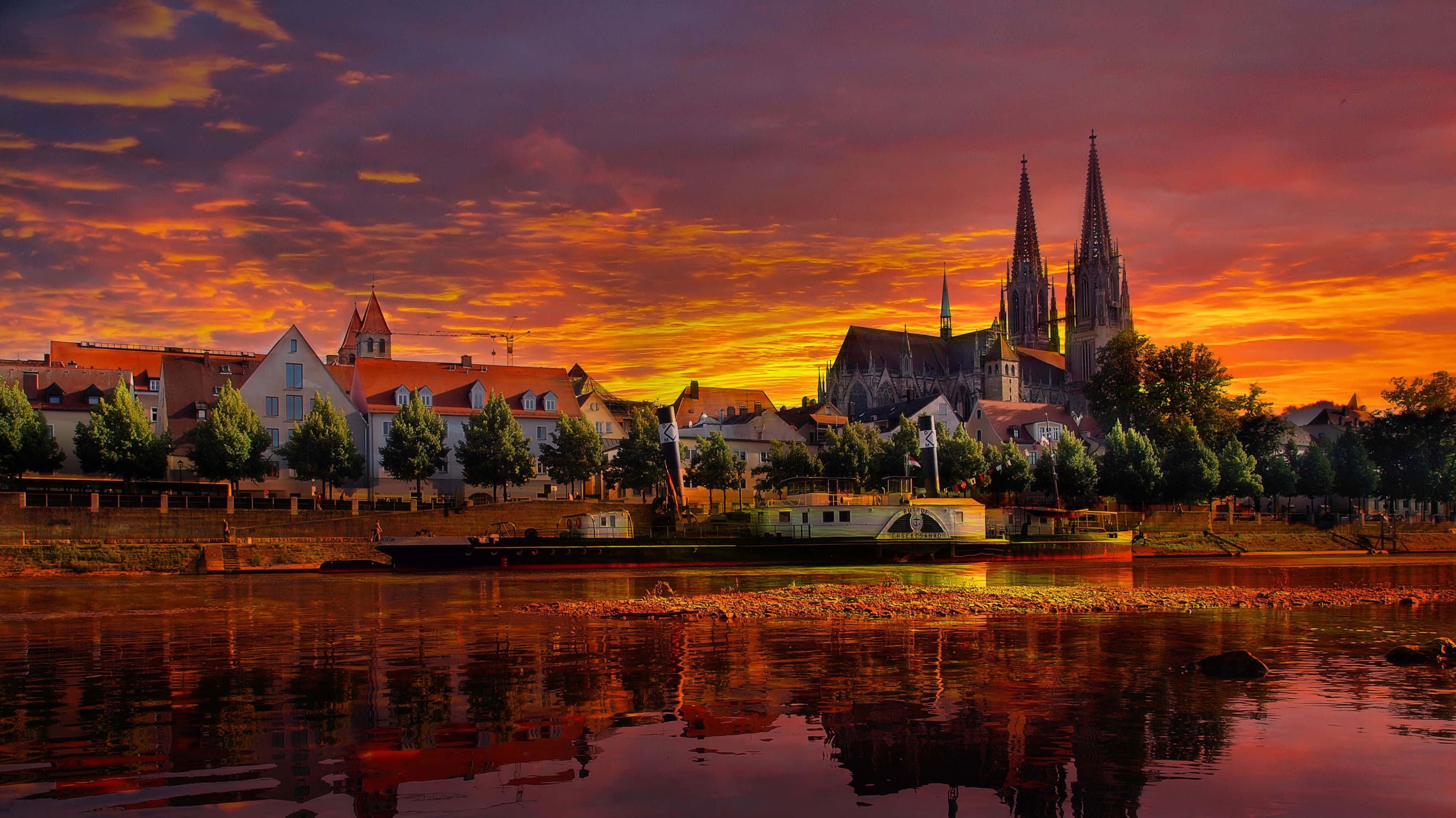 Germany 桌布 壁紙 — 在歷史悠久的河畔小鎮上空，夕陽以火熱的橙色和紫色調繪製天空，大教堂和船隻倒映在平靜的水面上。