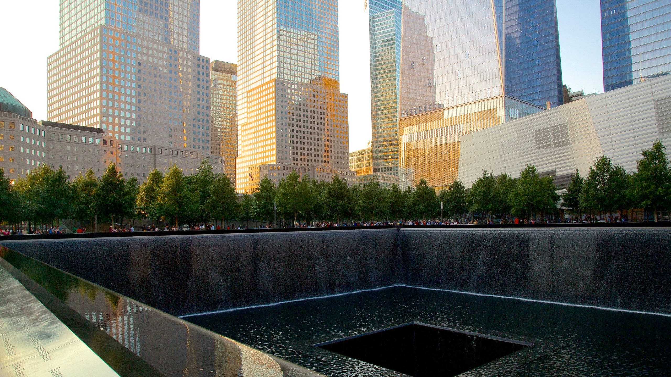 911 Memorial 桌布 壁紙 — 在歸零地，一個莊嚴的倒影池被高聳的玻璃摩天大樓環繞，沐浴在金色的日落光芒中。