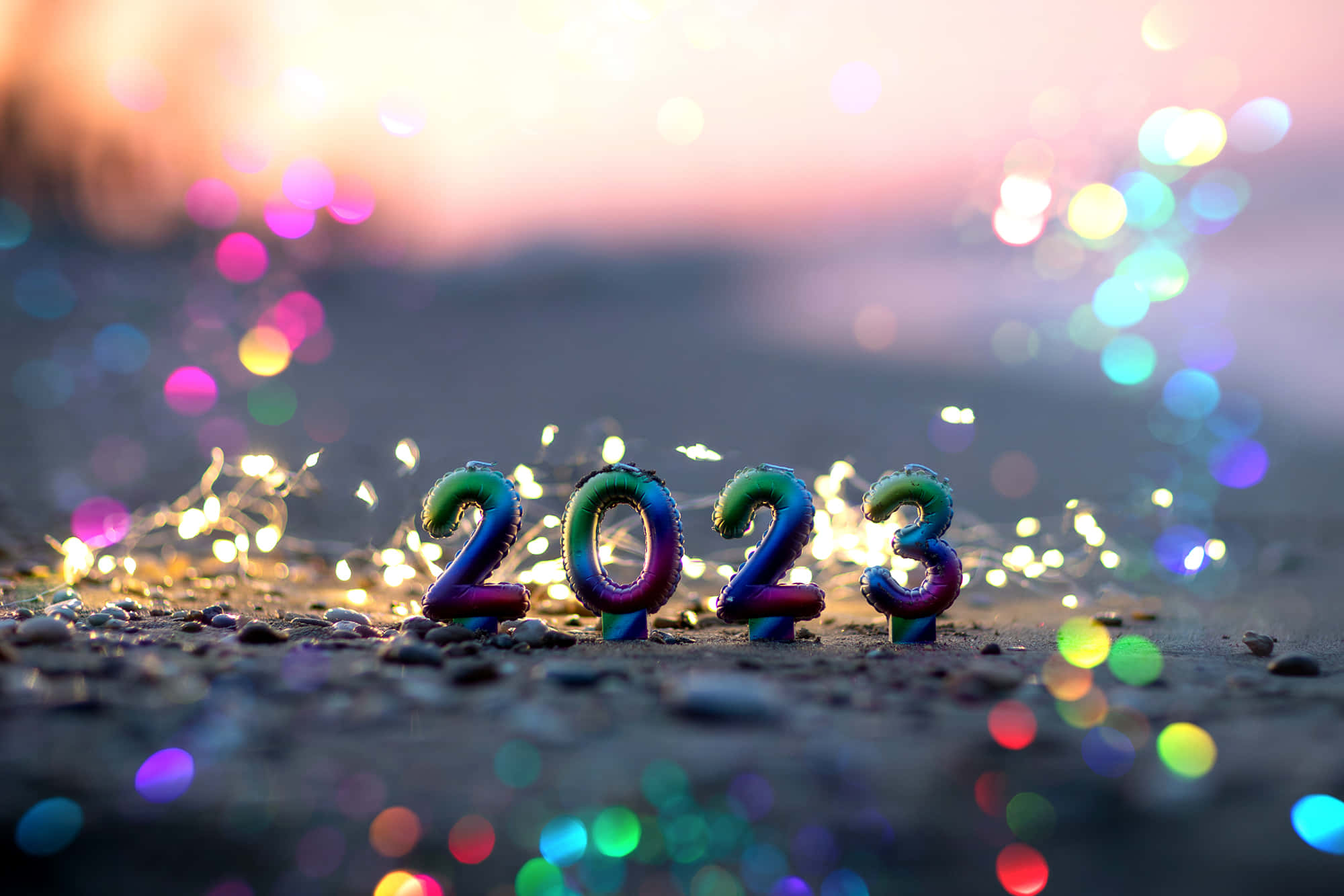 新年動畫 桌布 壁紙 — 在沙灘上閃爍燈光和日落散景背景下的2023年彩虹色氣球數字。