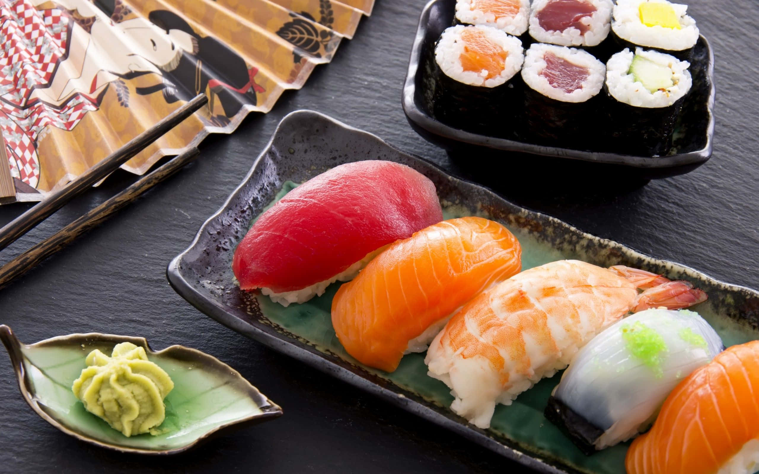 Sushi 桌布 壁紙 — 在深色石板表面上，新鮮壽司的鮮豔組合，包括吞拿魚、三文魚和蝦握壽司，以及卷壽司和芥末。