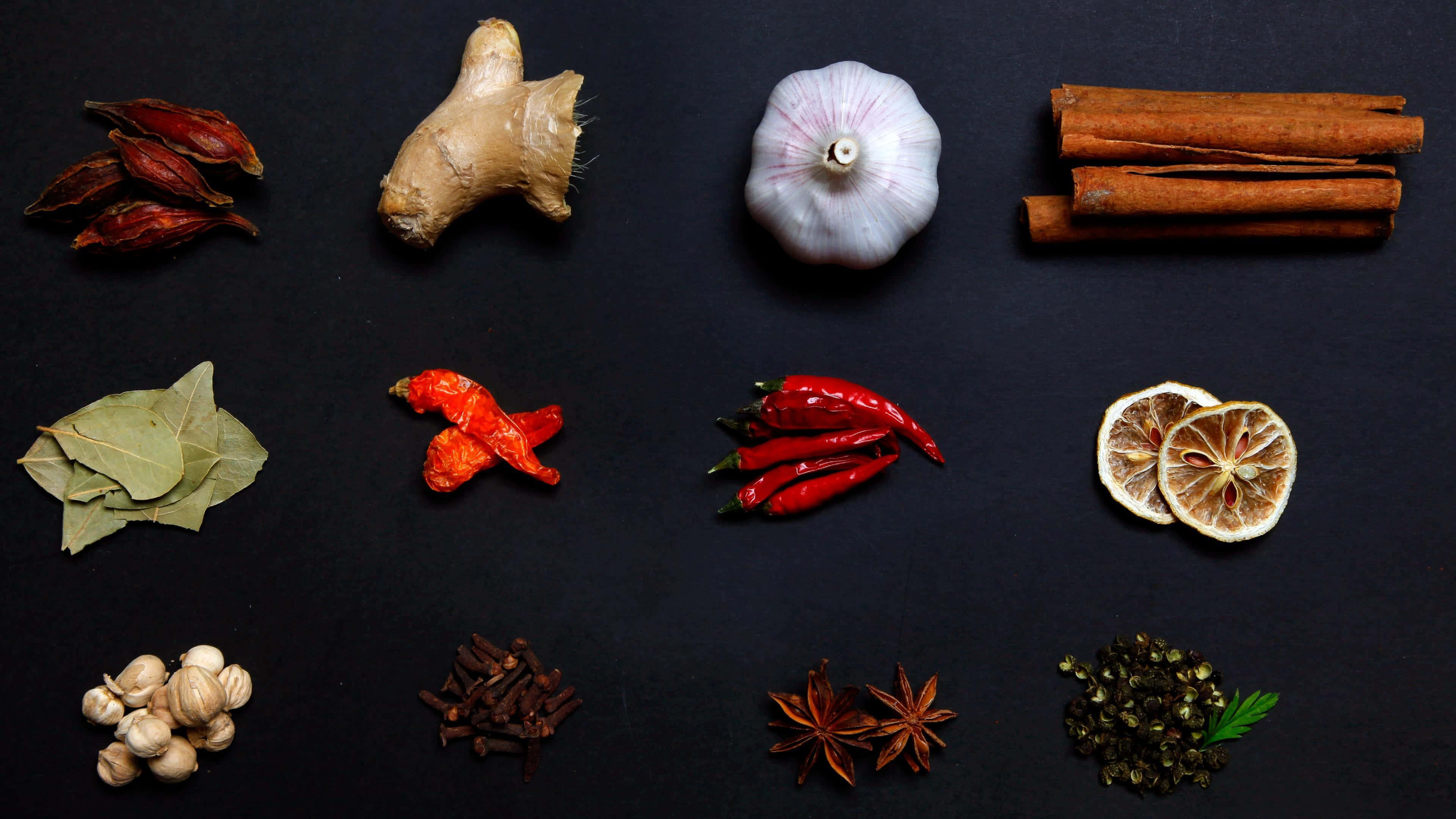 Spices 桌布 壁紙 — 在深色表面上平鋪擺放色彩鮮豔的香料和香草，包括肉桂棒、大蒜、生薑和乾辣椒。