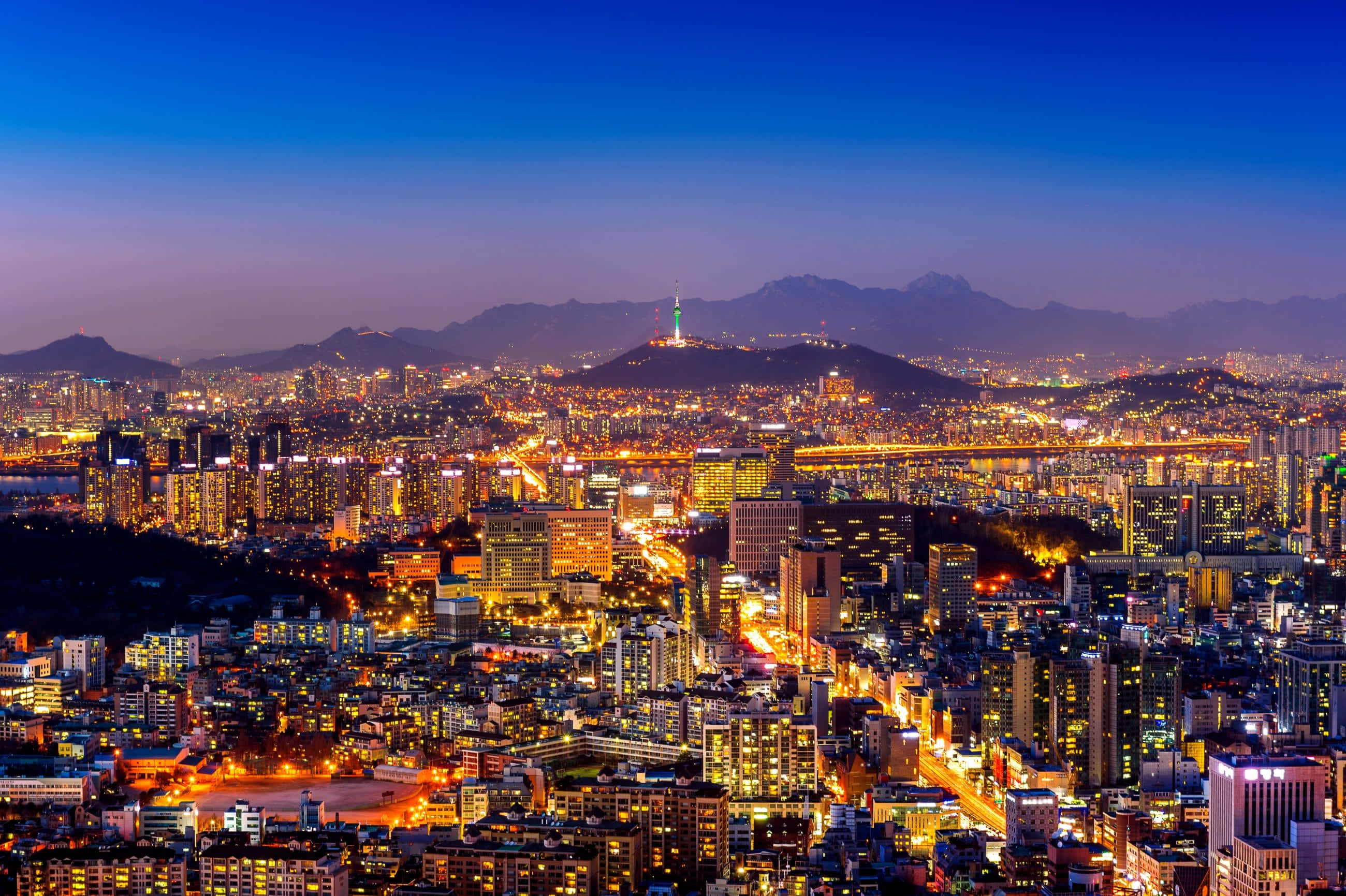 South Korea 桌布 壁紙 — 在深藍色天空下，暮色中廣闊的城市景觀，閃爍的摩天大樓和遠處的山脈。