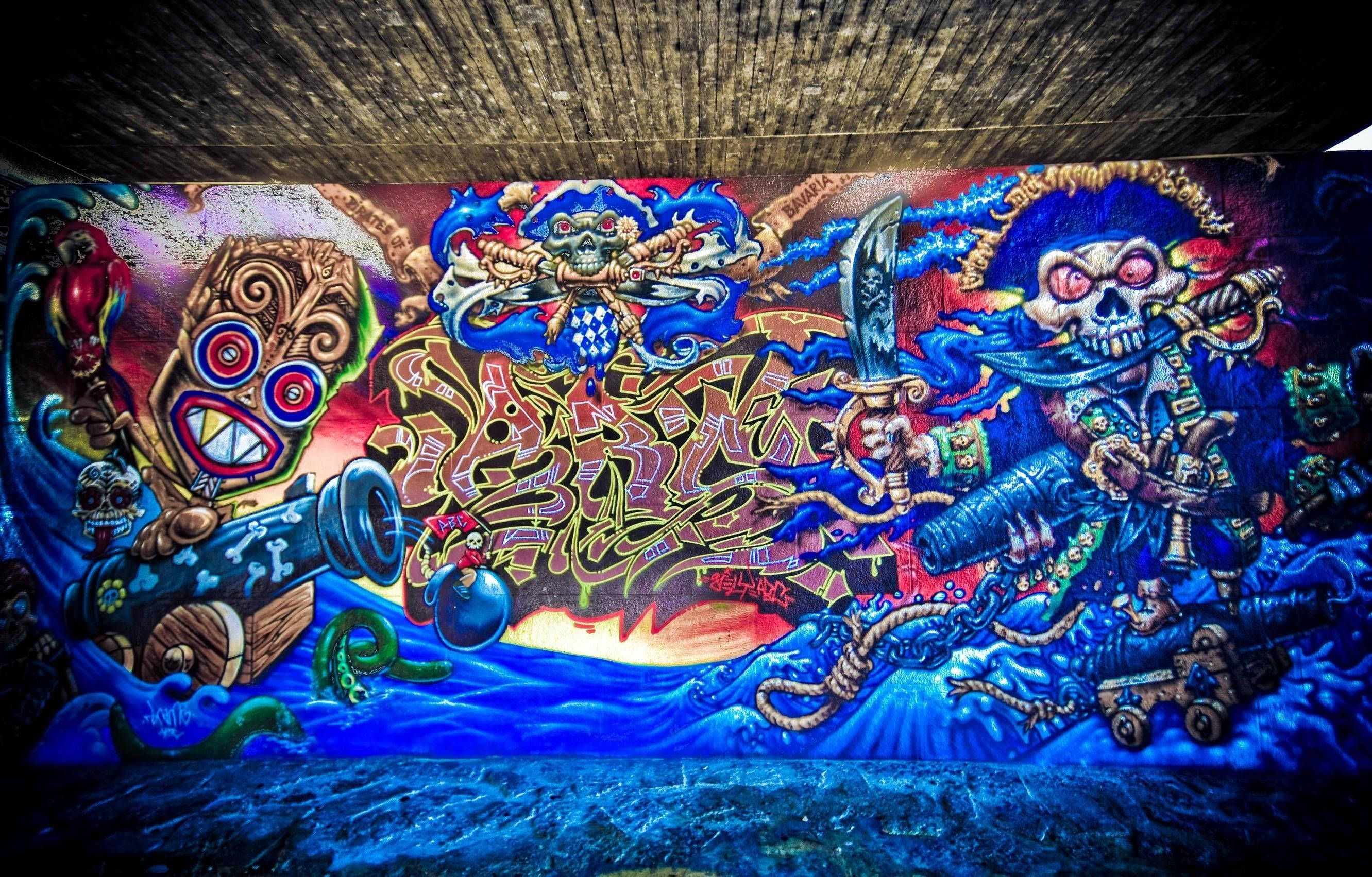 塗鴉 桌布 壁紙 — 在混凝土天橋下，充滿活力的海盜骷髏頭和大砲塗鴉壁畫，以藍色、紅色和金色爆發。