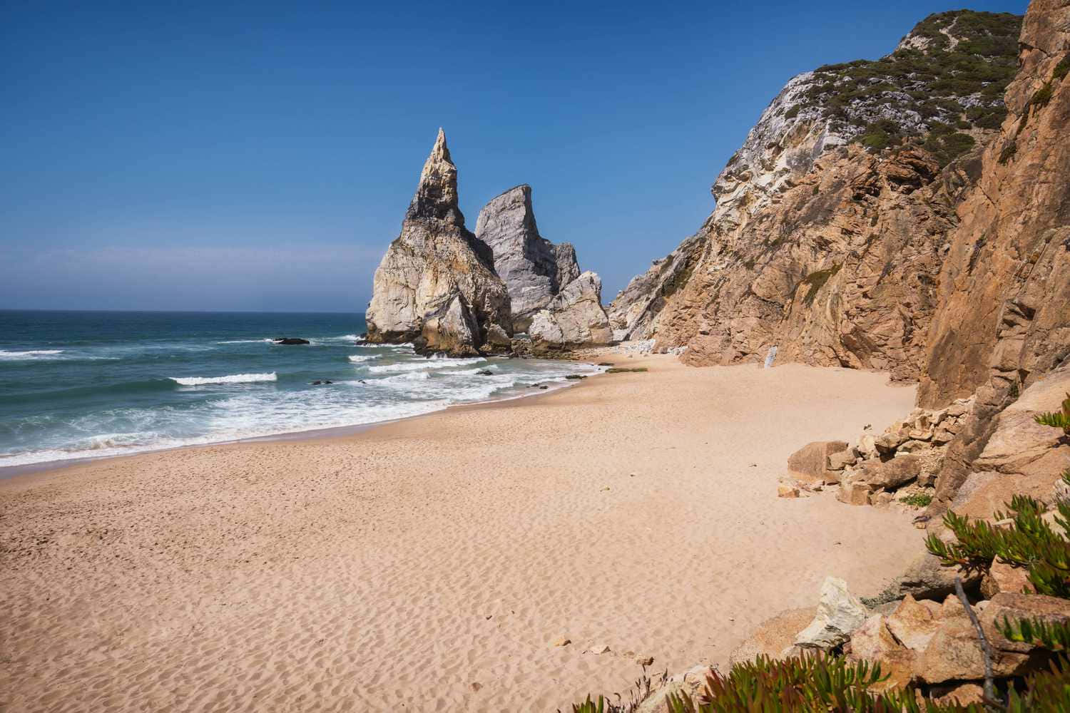 Portuguese Beach 桌布 壁紙 — 在清澈的藍天下，一個寧靜的沙灘在 towering 岩石懸崖旁蜿蜒，兩個突出的海蝕柱從碧綠的海水中升起。