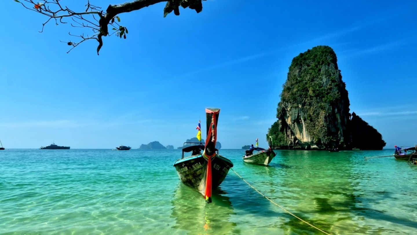 Thailand 桌布 壁紙 — 在清澈的藍天下，充滿活力的碧綠海水環繞著高聳的叢林覆蓋的石灰岩島嶼附近的傳統長尾船。