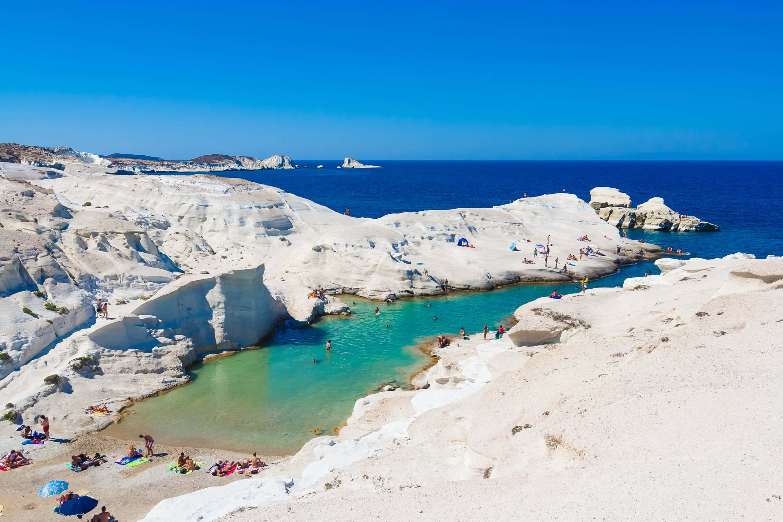Greek Beach 桌布 壁紙 — 在清澈的藍天下，白色石灰岩懸崖中鑲嵌著充滿活力的藍綠色潟湖，海灘遊客正在放鬆。