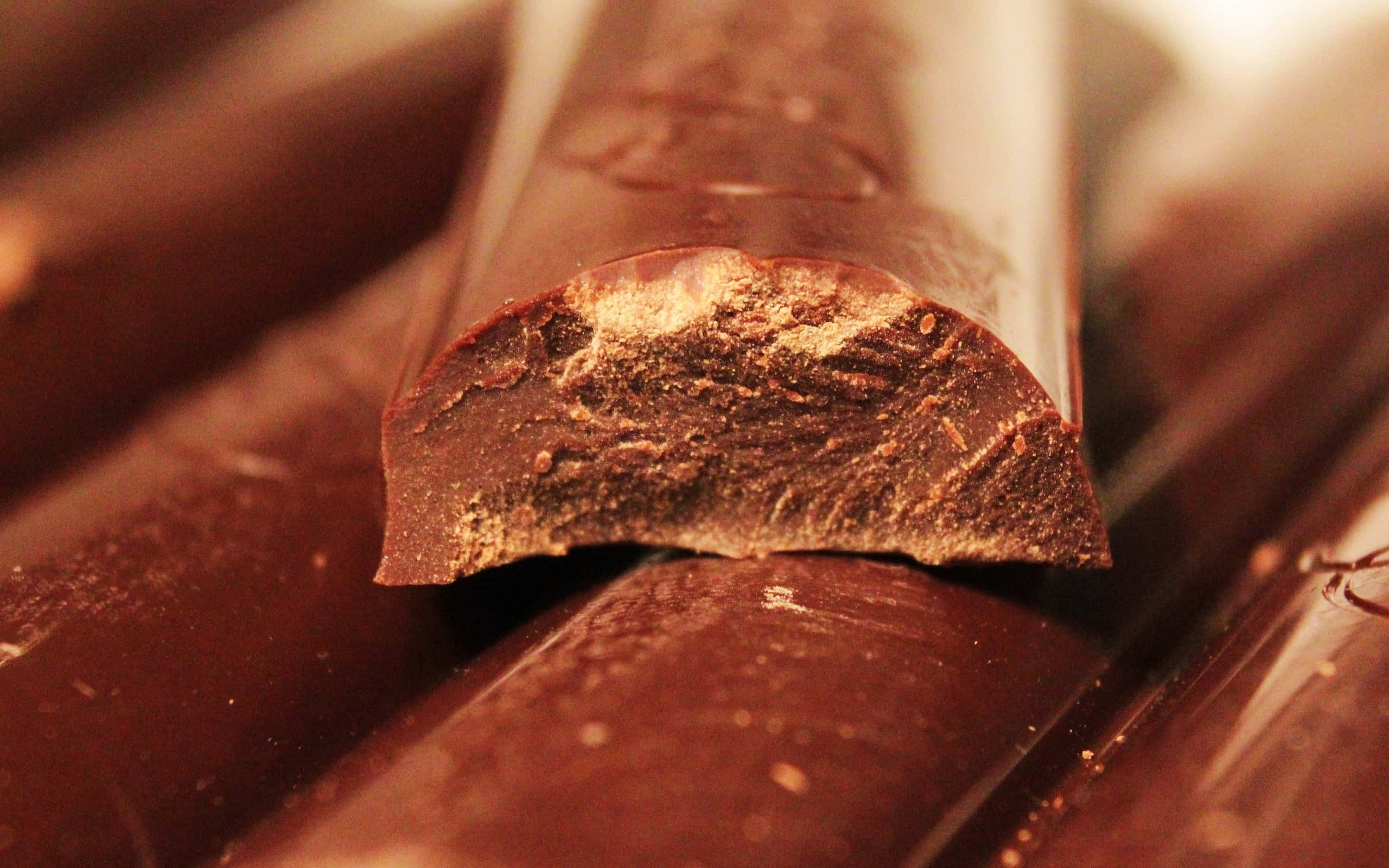 Brown Chocolate 桌布 壁紙 — 在溫暖燈光下，特寫一塊破碎的黑朱古力棒，展現其紋理豐富的內部。
