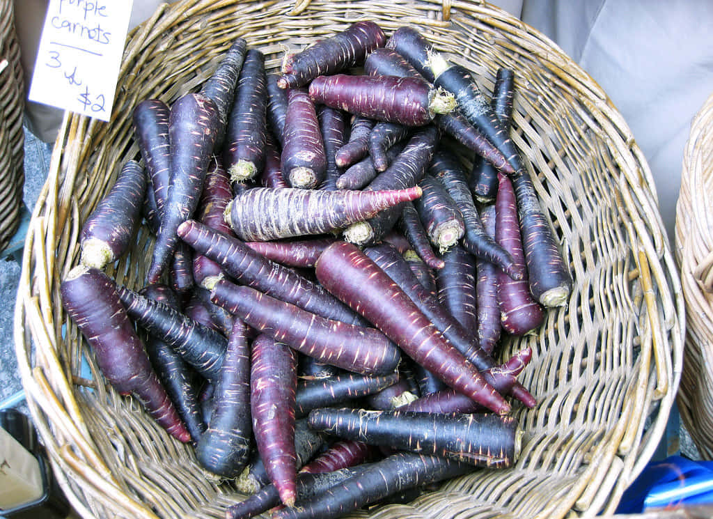 Purple Carrots 桌布 壁紙 — 在熙熙攘攘的農夫市場攤位上，一個裝滿鮮豔紫色胡蘿蔔的籃子。