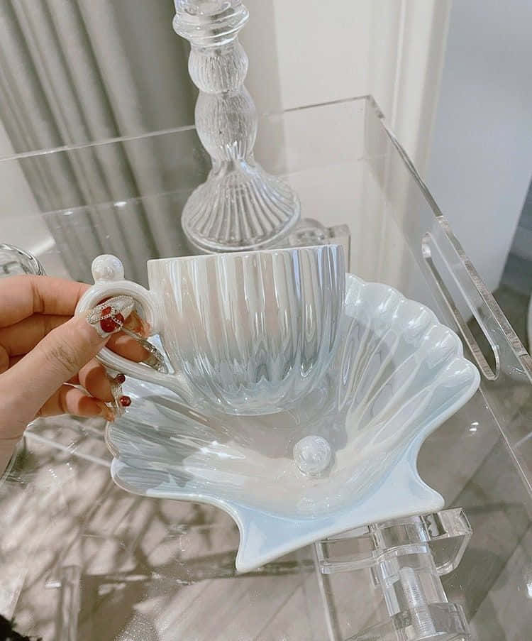 在理想園林中品味此刻 壁紙 — 一隻手拿著一個精緻的棱紋玻璃杯，放在一個扇貝形白色碟子上，置於透明亞加力桌上，背景有一個玻璃燭台。