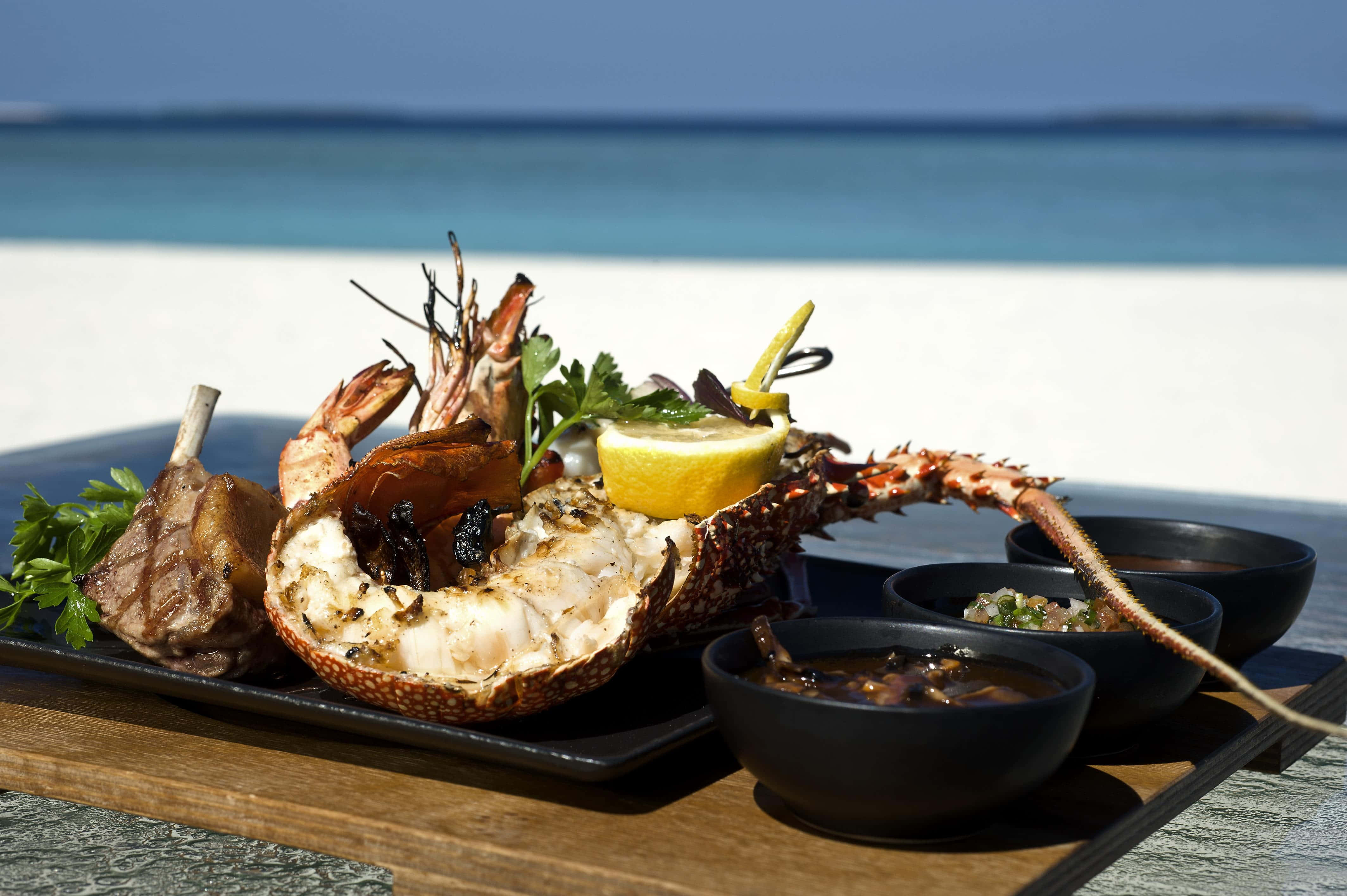 Beach Picnic 桌布 壁紙 — 在碧綠海洋和白色沙灘的背景下，烤龍蝦配檸檬和香草，放在托盤上，旁邊有蘸醬。