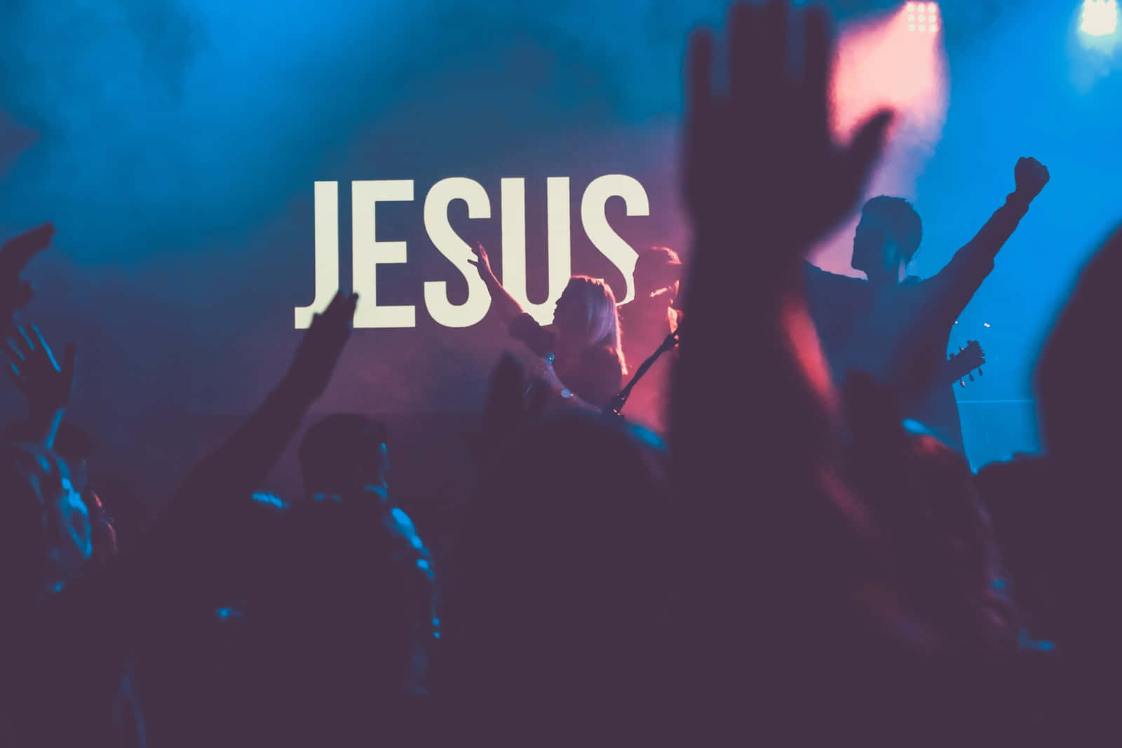 舉手的人群在藍色粉紅色燈光下敬拜，帶有「JESUS」文字。