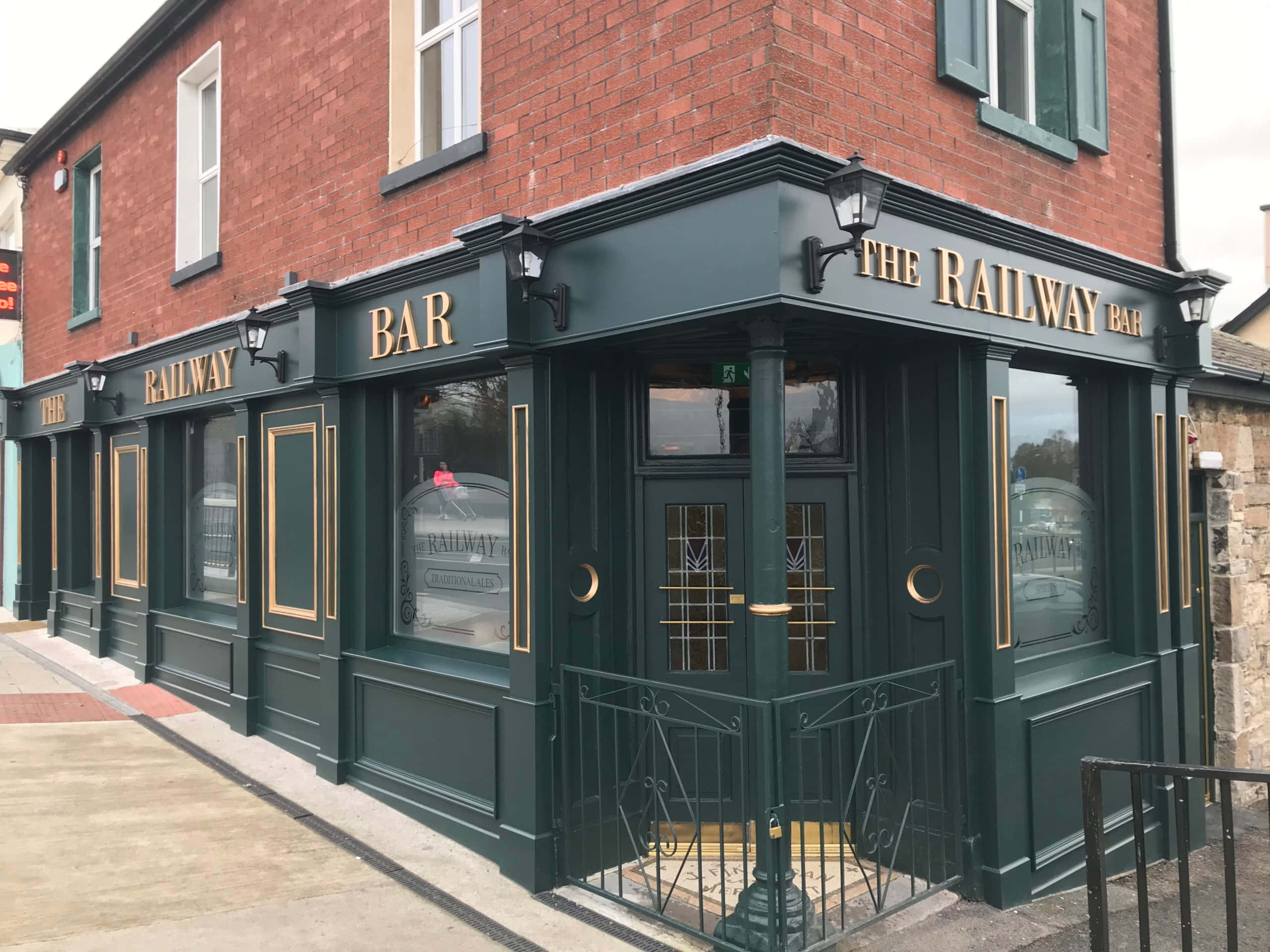 在私家酒吧中悠閒品啤酒 壁紙 — 一間名為「The Railway Bar」的經典磚砌酒吧，帶有深綠色飾邊和金色字樣，矗立在陰天街道的角落。