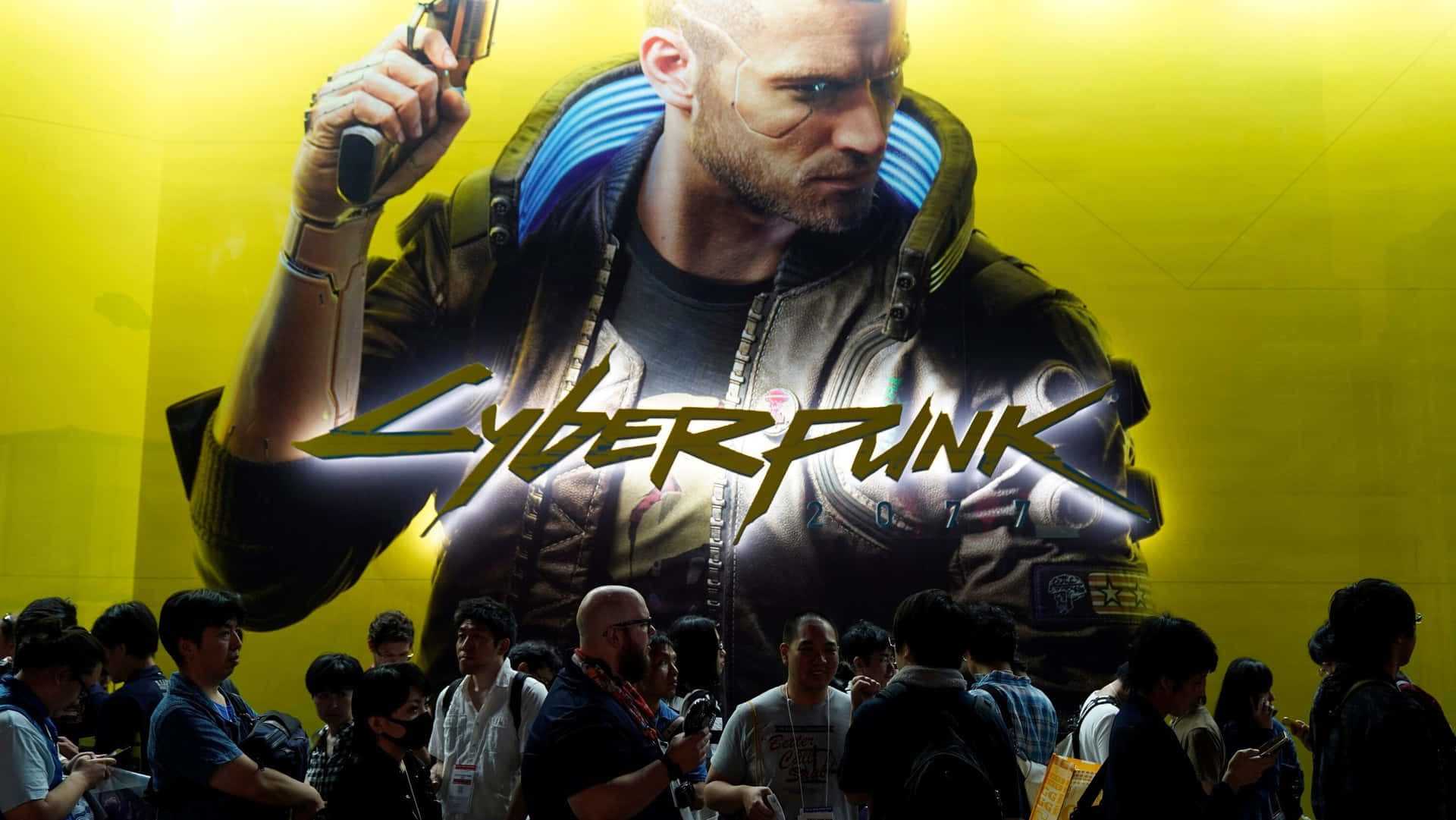 人群觀看一個大型 Cyberpunk 2077 宣傳海報，上面有一個機械人拿著槍。