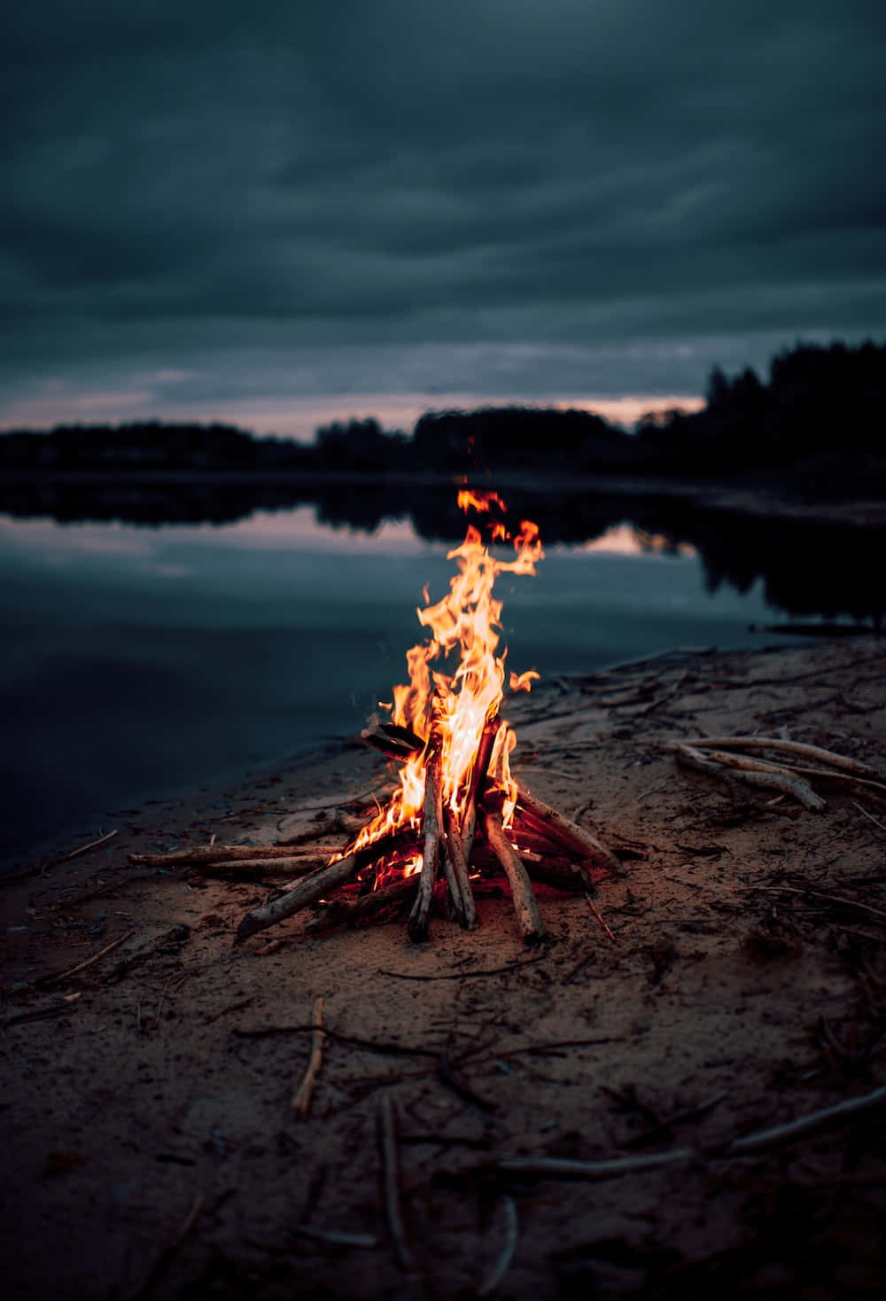 在篝火旁度過寧靜舒適的夜晚 壁紙 — 一個充滿活力的營火在沙灘湖岸上噼啪作響，下方是陰沉的暮色天空，烏雲密佈，遠處的樹木倒映在平靜的水面上。