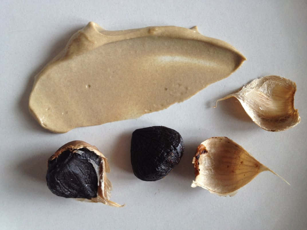 Black Garlic 桌布 壁紙 — 在純白表面上，烤蒜瓣和紙狀蒜皮旁邊有一抹奶油色米色醬料。