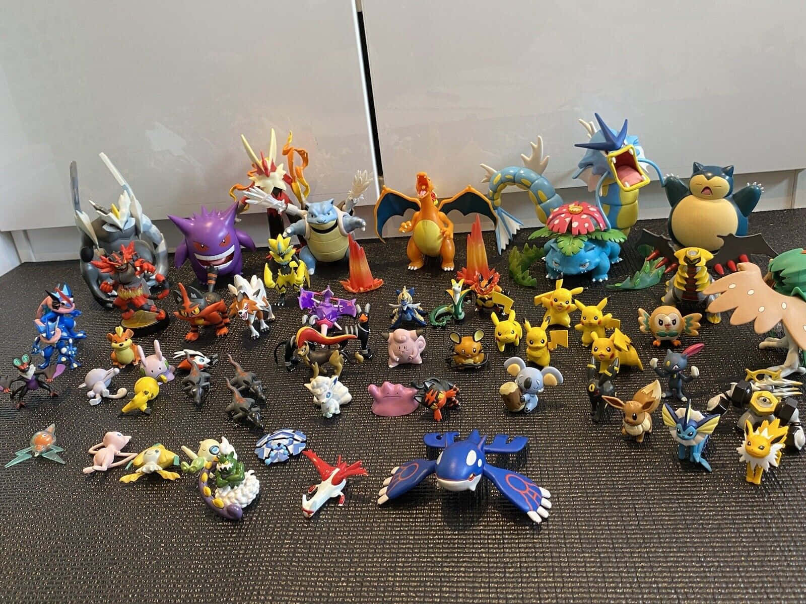 Pokemon Figures 桌布 壁紙 — 在素色牆壁前，暗色紋理表面上排列著色彩繽紛的寵物小精靈公仔收藏。