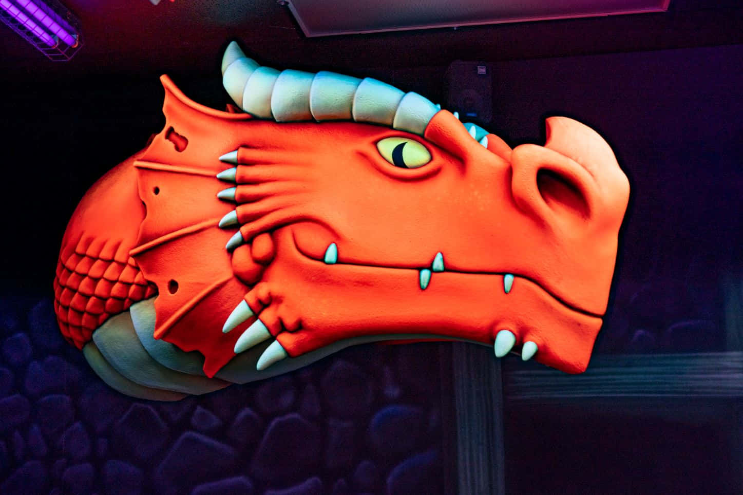 Dragons 桌布 壁紙 — 在紫色燈光下，一個鮮豔的橙色龍頭雕塑，帶有發光的綠色眼睛和白色角，安裝在深色紋理牆上。