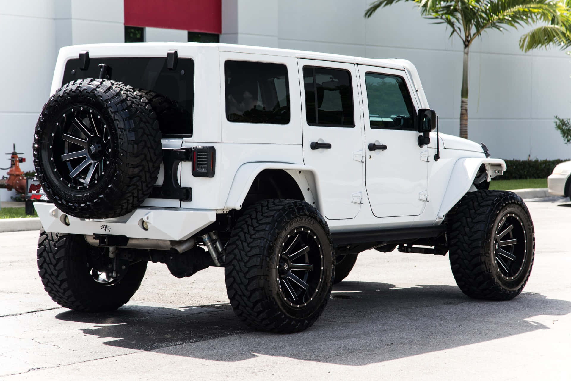 在經典Jeep Wrangler中探索戶外極限 壁紙 — 一輛完美無瑕的白色升高Jeep Wrangler，配備超大黑色輪胎和安裝的備用輪胎，停放在陽光明媚的混凝土場地上。