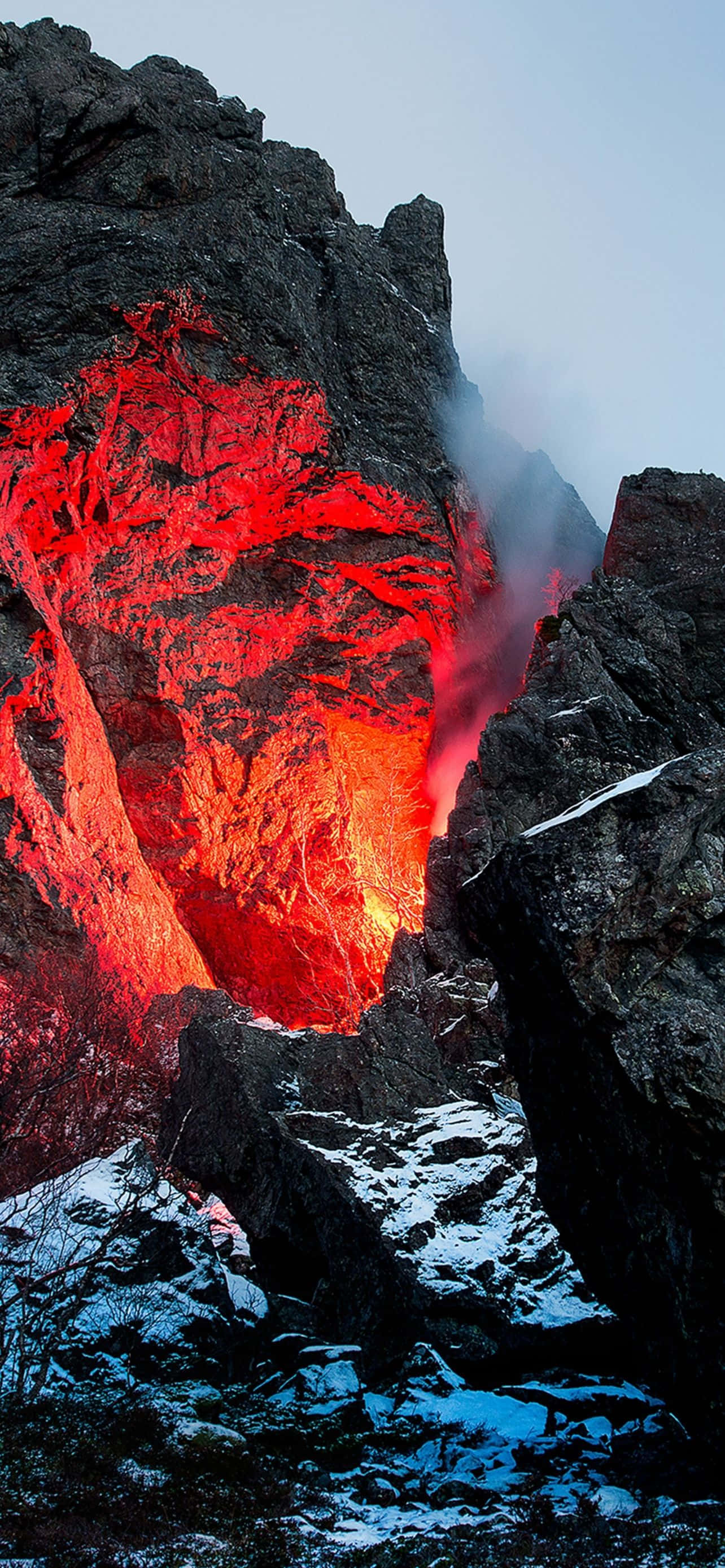 Mountain iPhone 桌布 壁紙 — 在蒼白的天空下，一道火山裂縫噴發出熾熱的紅色熔岩，落在覆蓋著白雪的岩石上。
