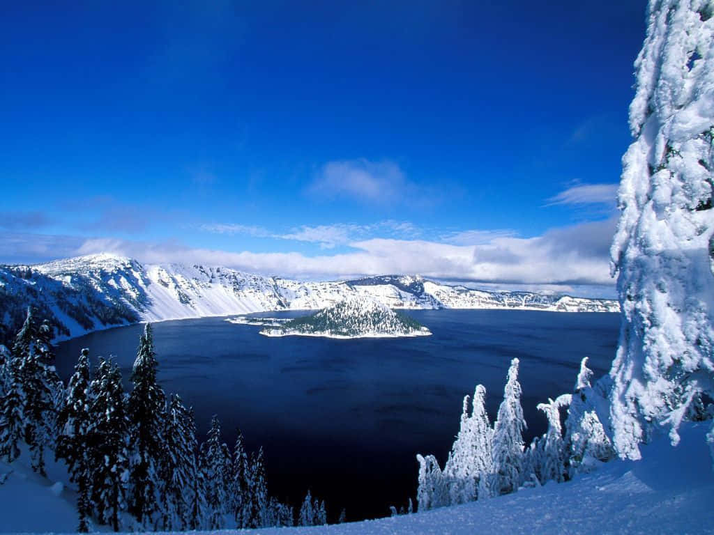 Frozen Lake 桌布 壁紙 — 在蔚藍天空下，一個被白雪覆蓋的火山口湖， fluffy 雲朵和掛滿霜的松樹框住了這片景色。