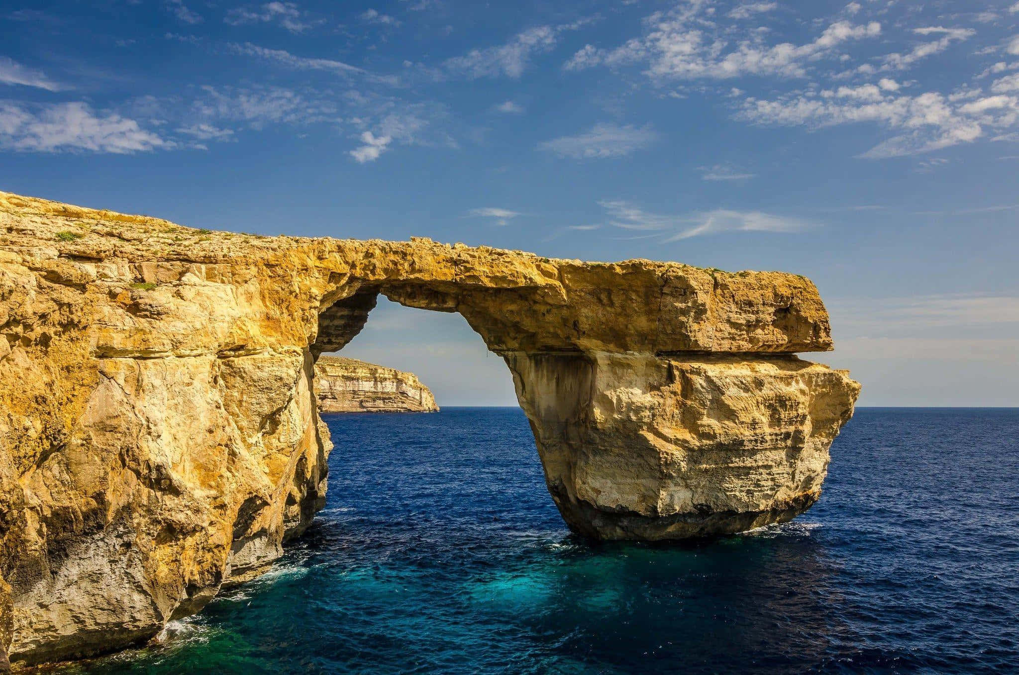 Malta 桌布 壁紙 — 在藍天白雲下，一道雄偉的金色岩石拱門從碧綠的海水中升起。